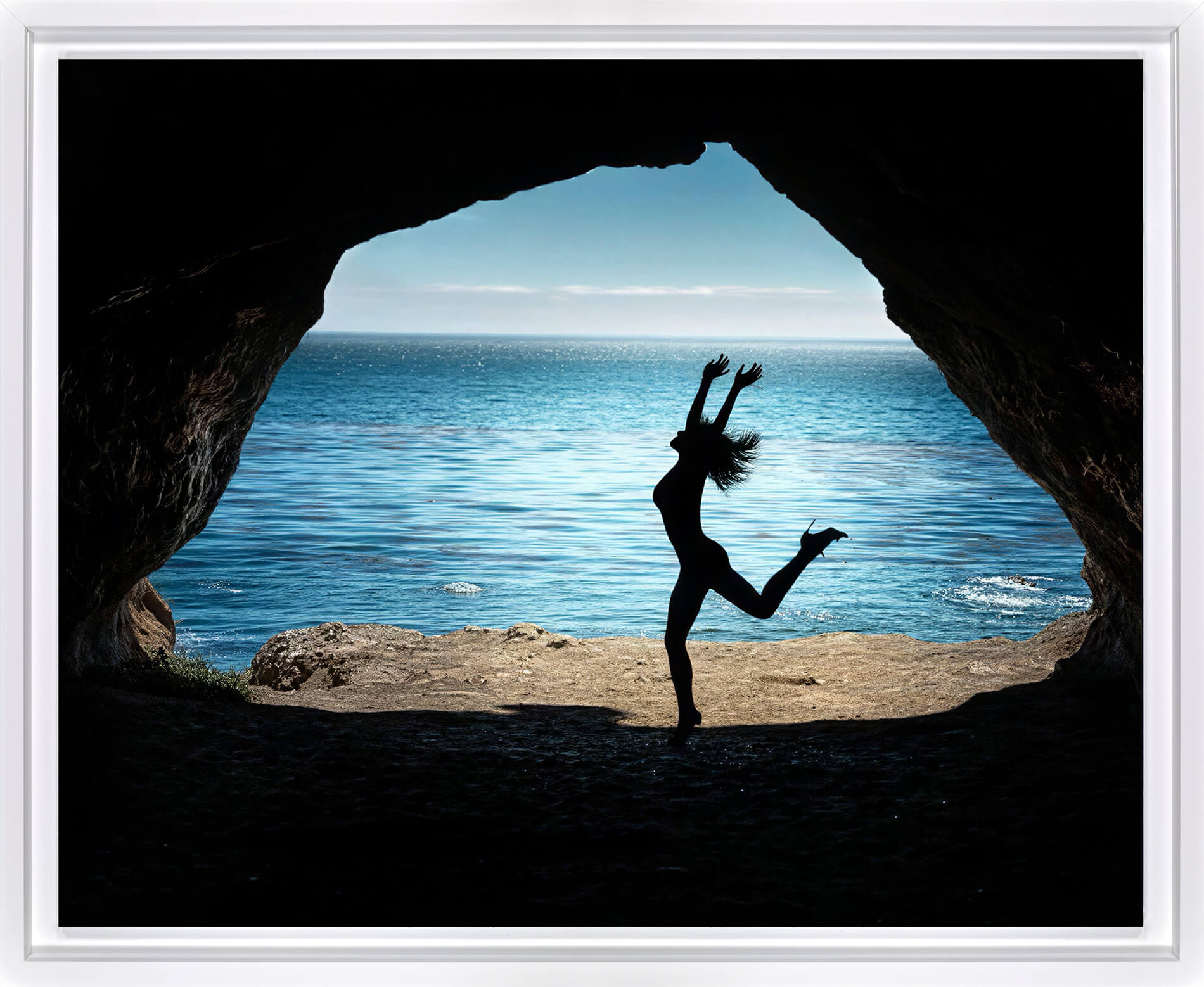 A framed photographic print showing a silhouetted dancer jumping in front of a cave opening that reveals a bright blue ocean and sky.