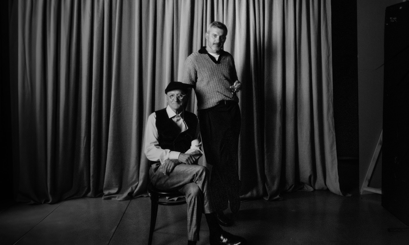 Two men posing in front of a curtain, one sitting and one standing.