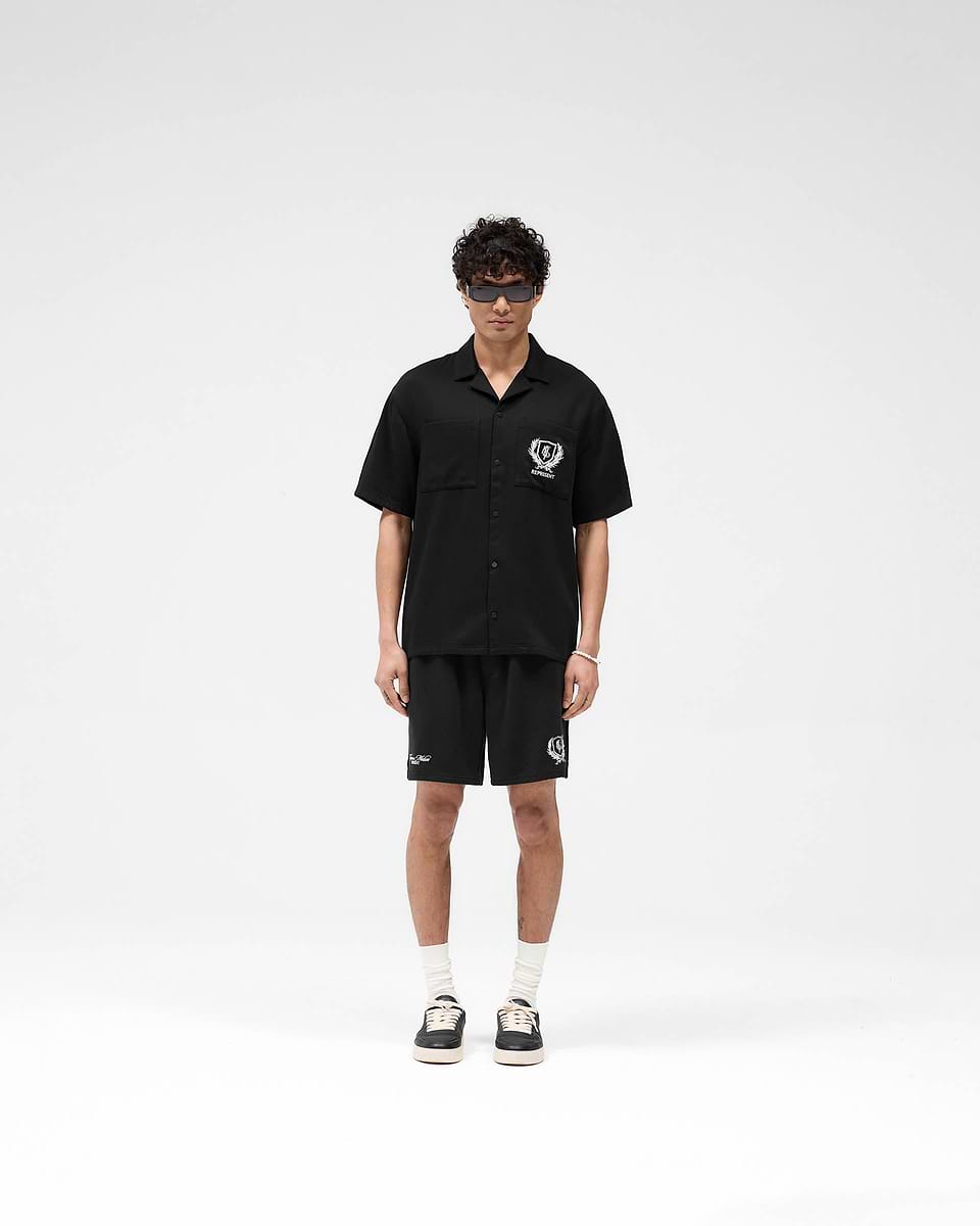 Man wearing a black short-sleeve shirt on a plain white background | Represent Crest Shirt in Jet Black | Shirt with chest pockets and embroidered graphics | SS25 Shirts | Represent Clo
