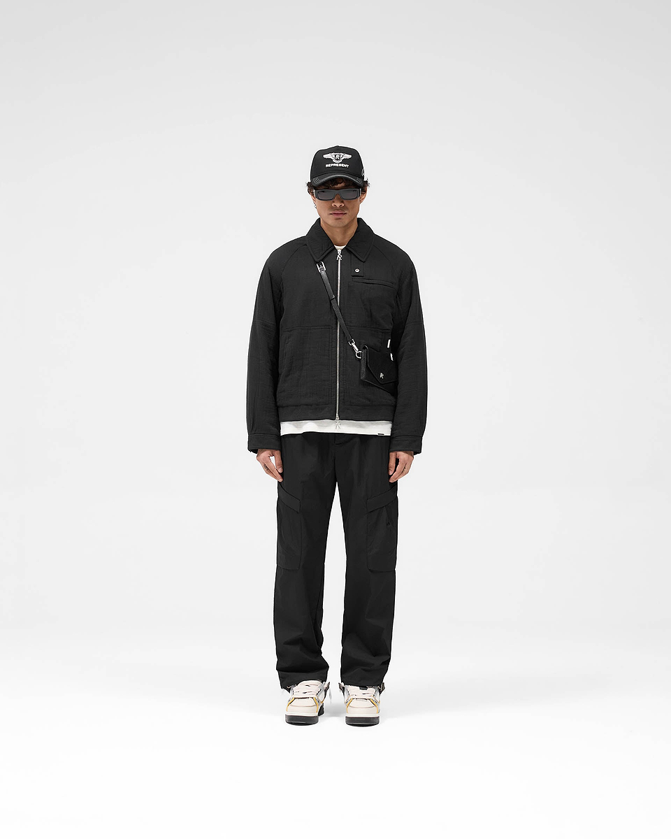 Man wearing black pants on a plain white background | Tech Cargo Pant in Jet Black | Lightweight pants with pleated cargo pockets and hybrid-style waistband | SS25 Pants | Represent Clo