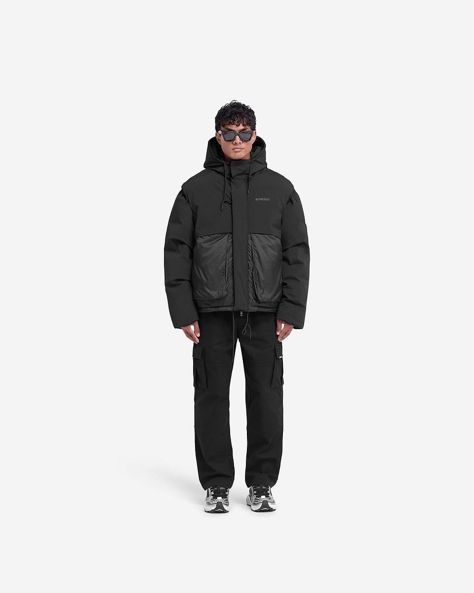 Man wearing a black puffer jacket on a plain white background | Layered Hooded Puffer in Jet Black | Duck down filled puffer with adjustable hood and tonal branding | FW24 Outerwear | Represent Clo