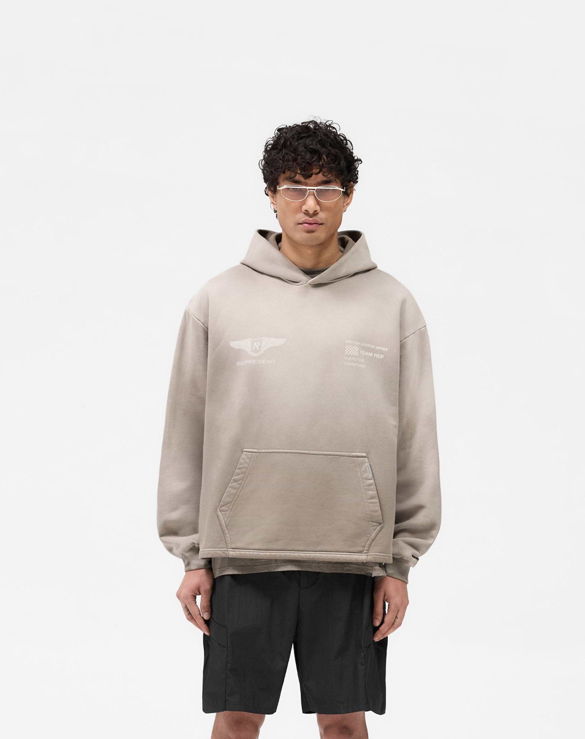 Man wearing an oversized dark taupe hoodie on a plain white background | Team Rep Hoodie in Dark Taupe | Heavy weight jersey cotton hoodie with vintage graphic | SS25 Hoodies | Represent Clo