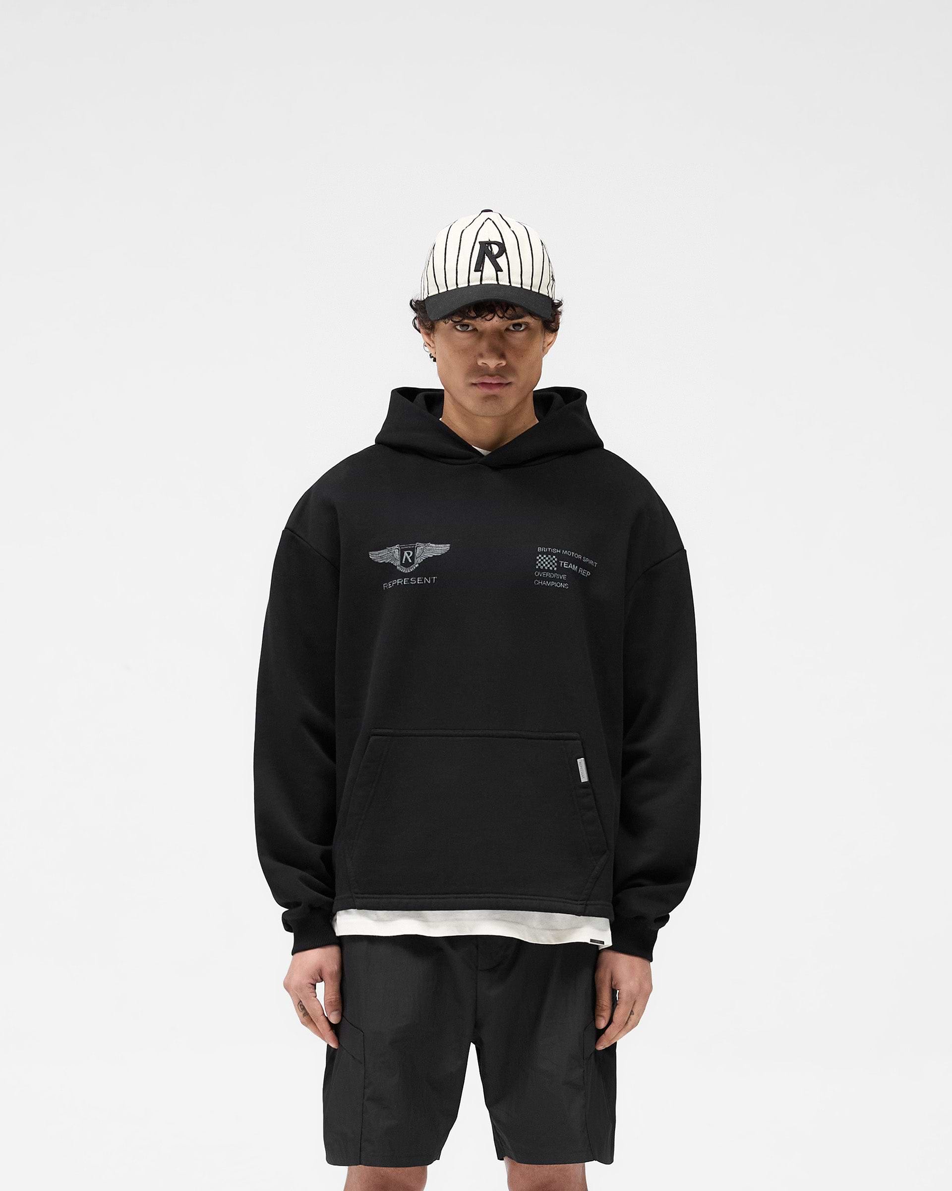 Man wearing an oversized black hoodie on a plain white background | Team Rep Hoodie in Black | Vintage worn graphic oversized hoodie | SS25 Hoodies | Represent Clo