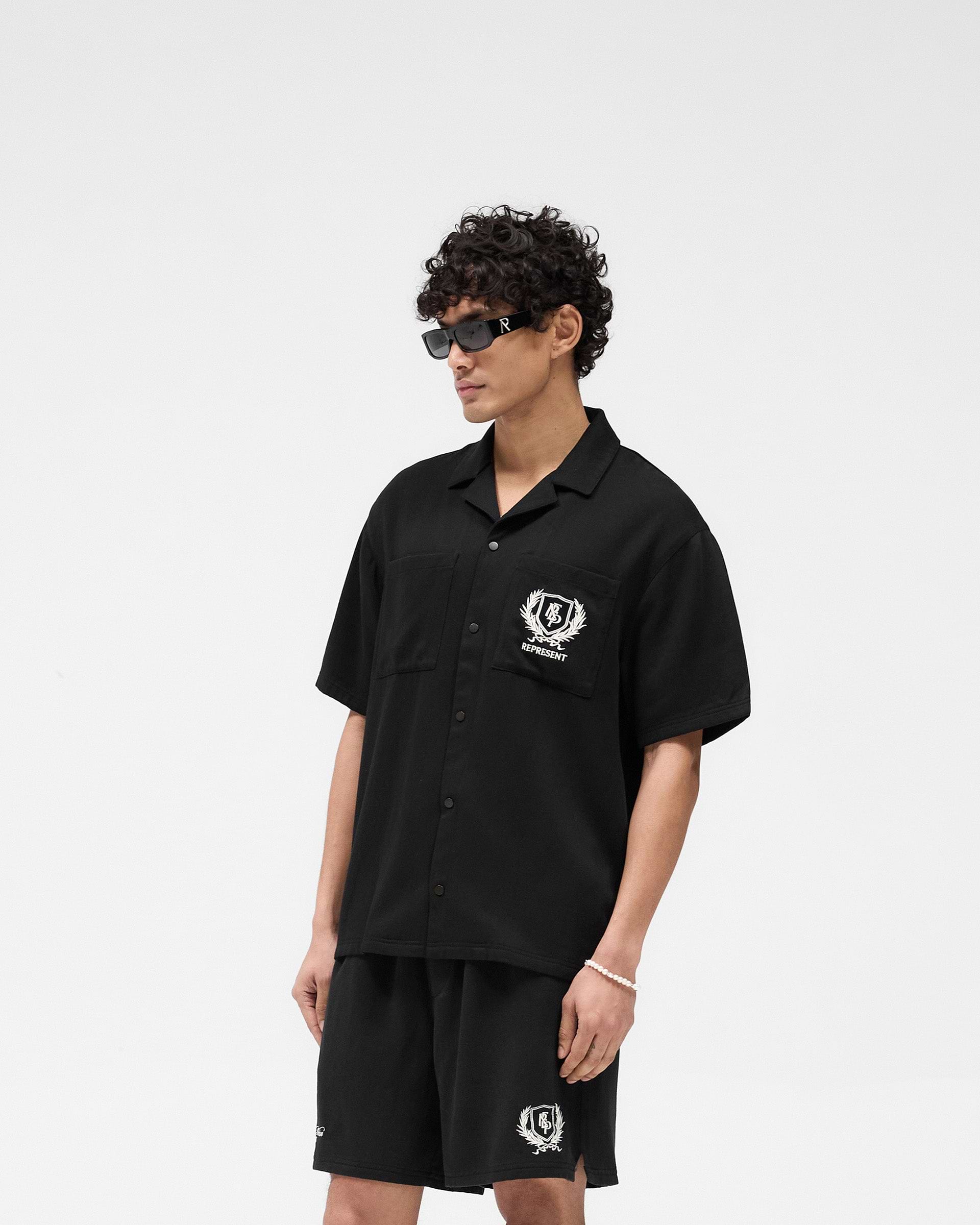 Man wearing a black short-sleeve shirt on a plain white background | Represent Crest Shirt in Jet Black | Lightweight shirt with embroidered crest and chest pockets | SS25 Shirts | Represent Clo