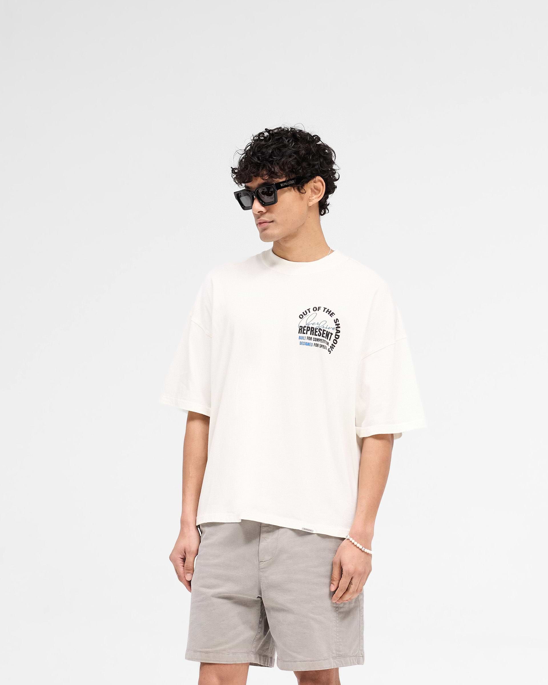 Man wearing a graphic t-shirt in white on a plain white background | Out Of The Shadows T-Shirt in Flat White | Boxy fit t-shirt with high build print | SS25 T-Shirts | Represent Clo