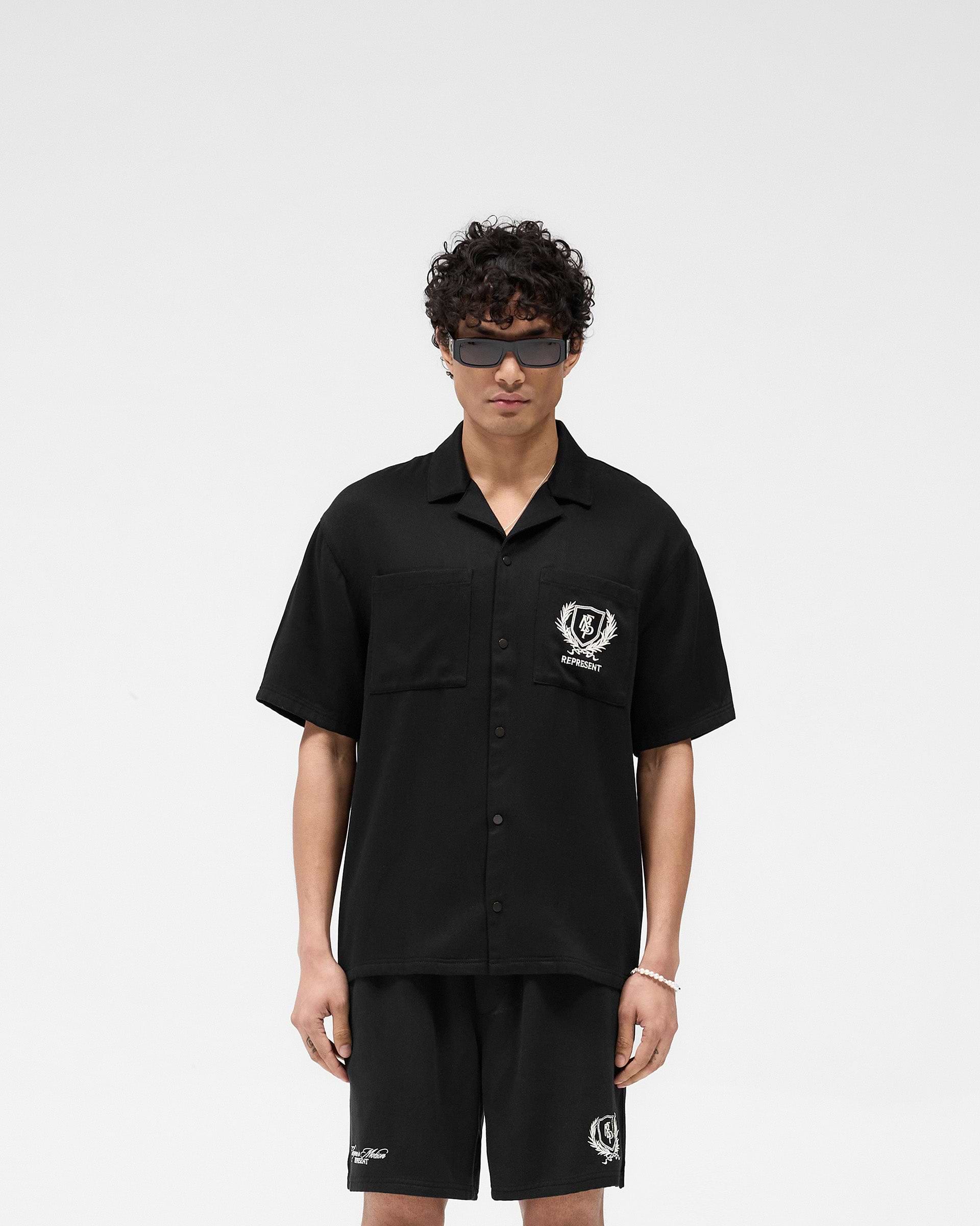 Man wearing a black short-sleeve shirt on a plain white background | Represent Crest Shirt in Jet Black | Lightweight shirt with dual chest pockets and back embroidery | SS25 Shirts | Represent Clo