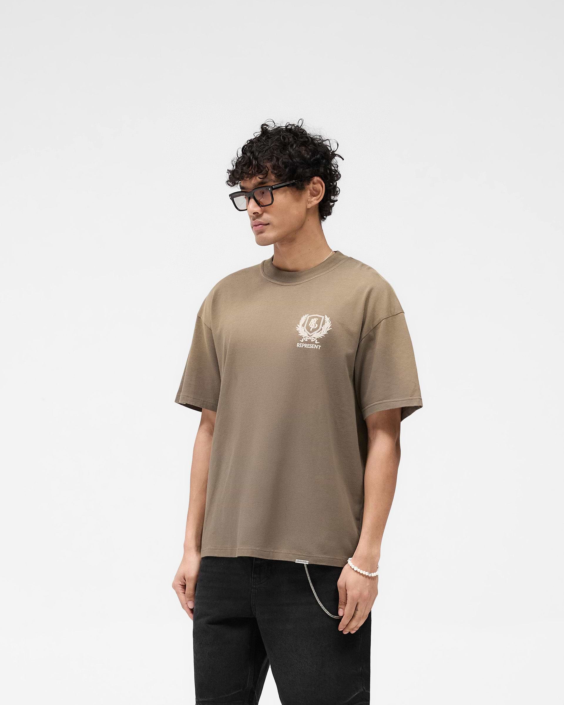 Man wearing a dark taupe graphic t-shirt on a plain white background | Crest T-Shirt in Dark Taupe | Oversized fit with crest and overdrive graphics | SS25 T-Shirts | Represent Clo
