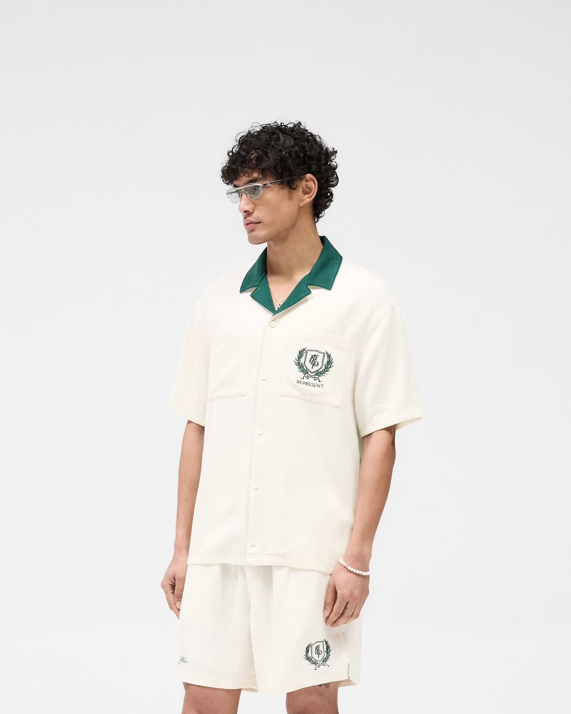Man wearing an antique white short-sleeve shirt on a plain white background | Represent Crest Shirt in Antique White | Short-sleeve shirt with contrast forest green collar | SS25 Shirts | Represent Clo