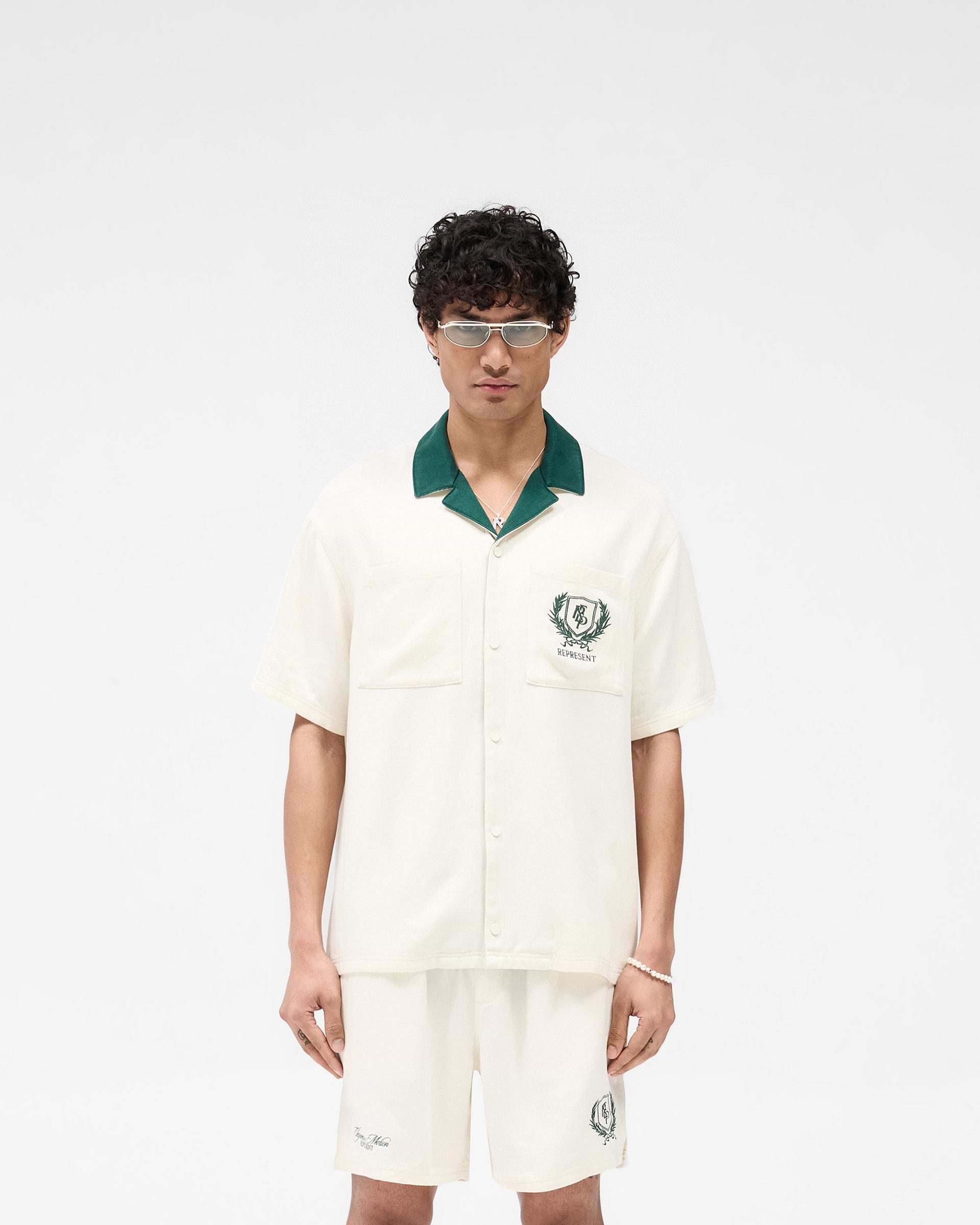 Man wearing an antique white short sleeve shirt with a forest green collar on a plain white background | Represent Crest Shirt in Antique White | Short sleeve shirt with dual chest pockets and embroidered back graphic | SS25 Shirts | Represent Clo
