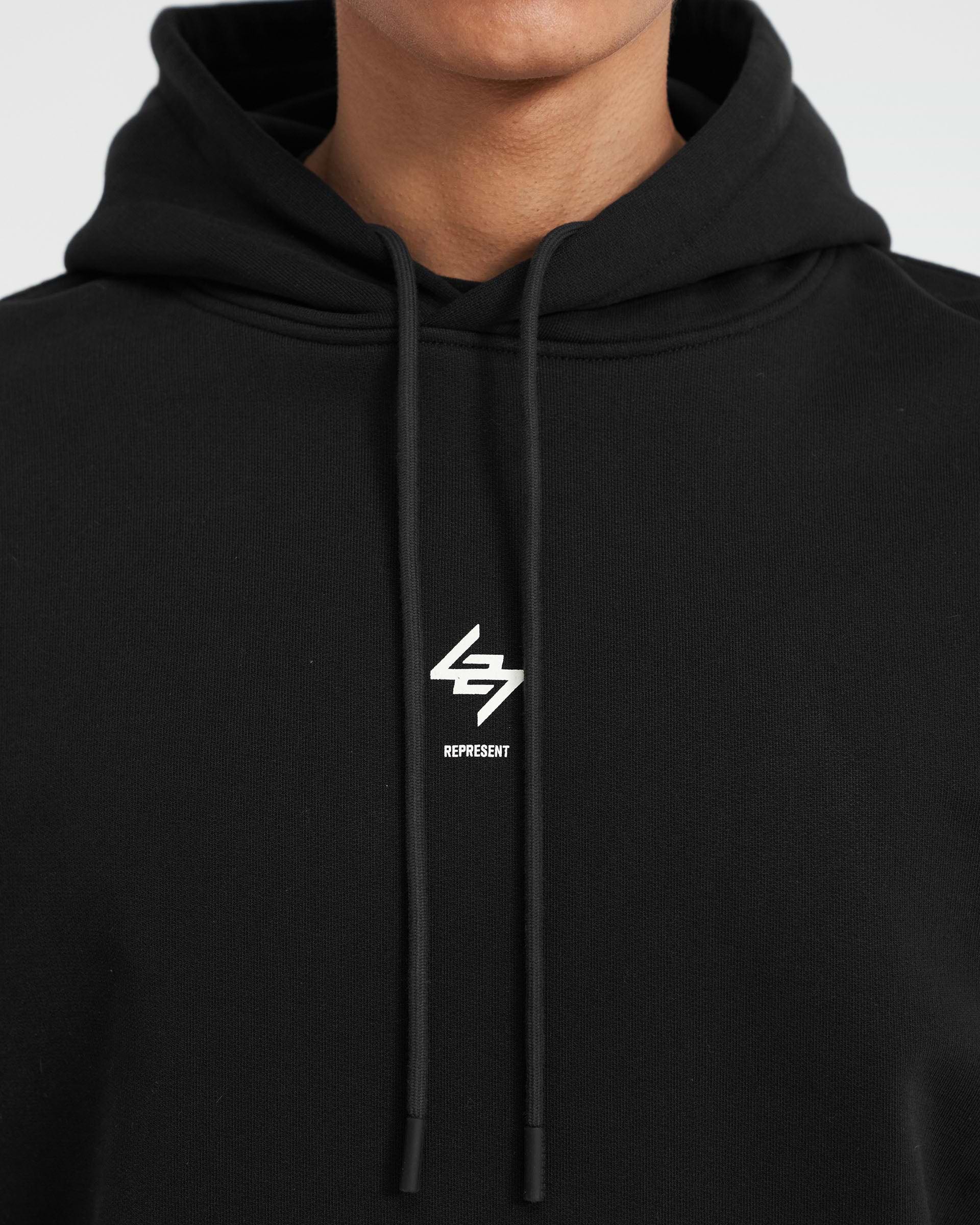 Man wearing an oversized black hoodie on a plain white background | 247 Oversized Hoodie in Jet Black | Hoodie with 247 branding and ribbed hems | 247 Hoodies | Represent Clo