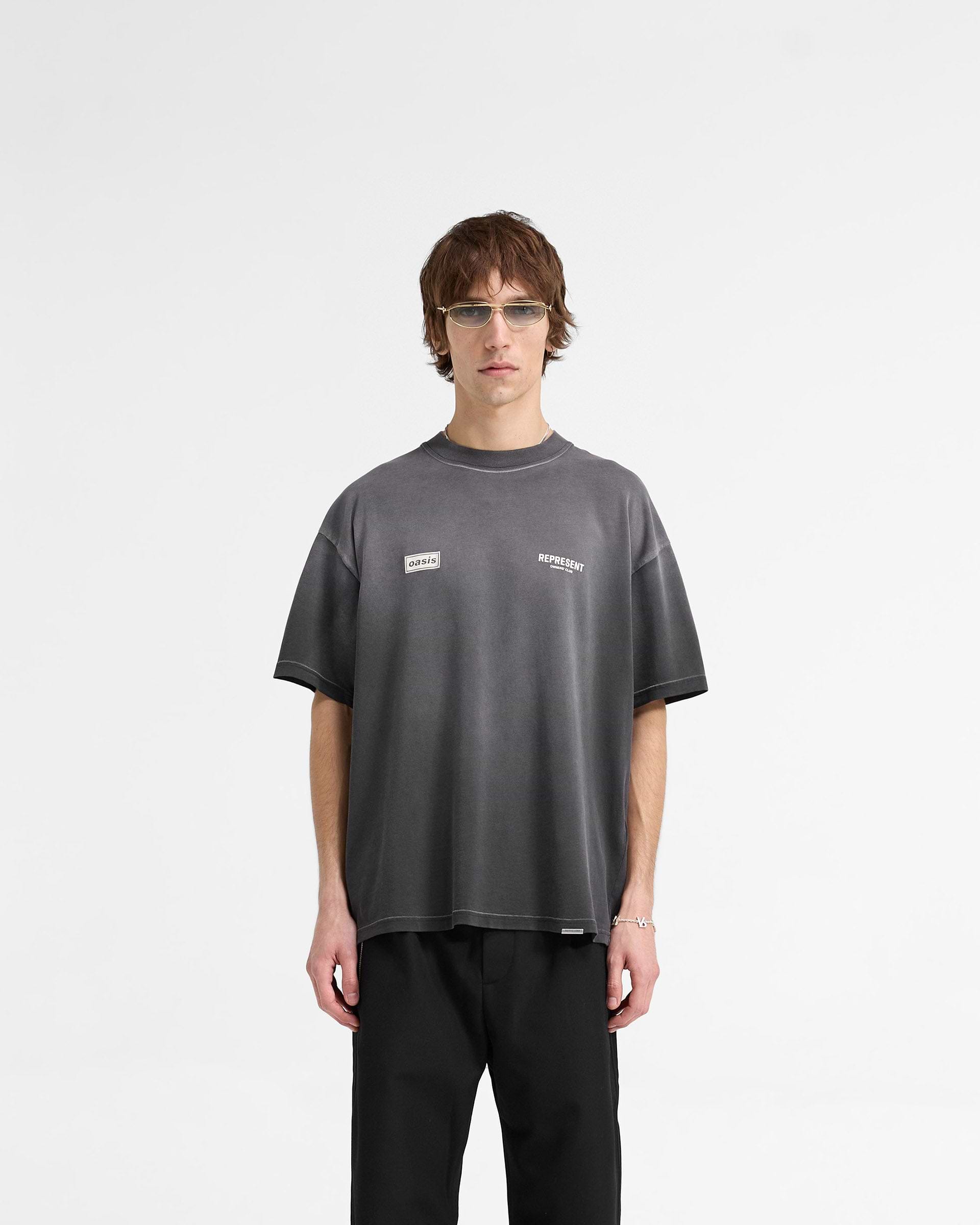 Man wearing an oversized black t-shirt on a plain white background | Represent X Oasis Owners Club T-Shirt in Stained Black | T-shirt with fly postered Oasis artwork and dual front logos | FW24 T-Shirts | Represent Clo