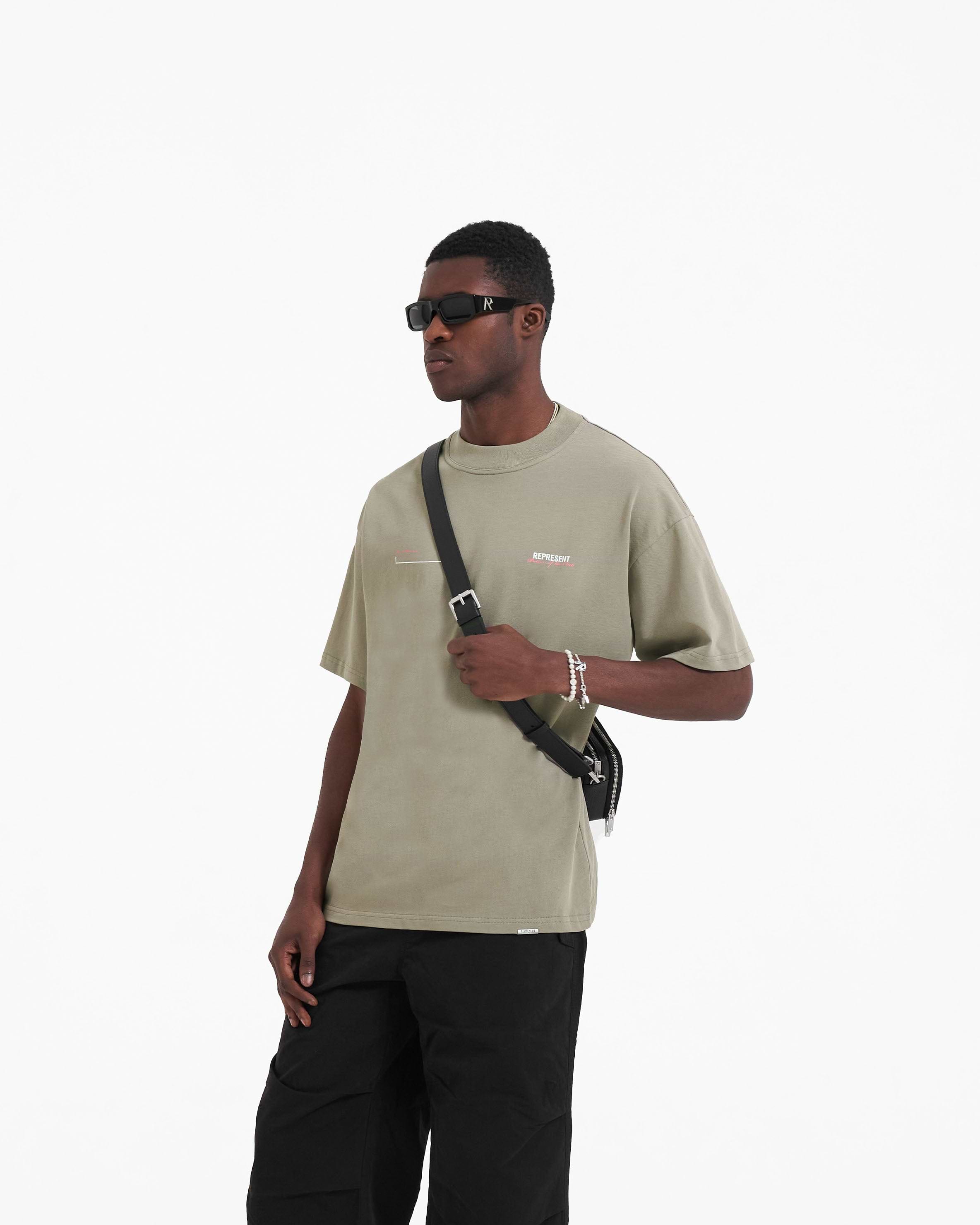 Man wearing a slightly oversized khaki t-shirt on a plain white background | Patron Of The Club T-Shirt in Khaki Pink | Oversized t-shirt with screen printed branding | Patrons T-Shirts | Represent Clo