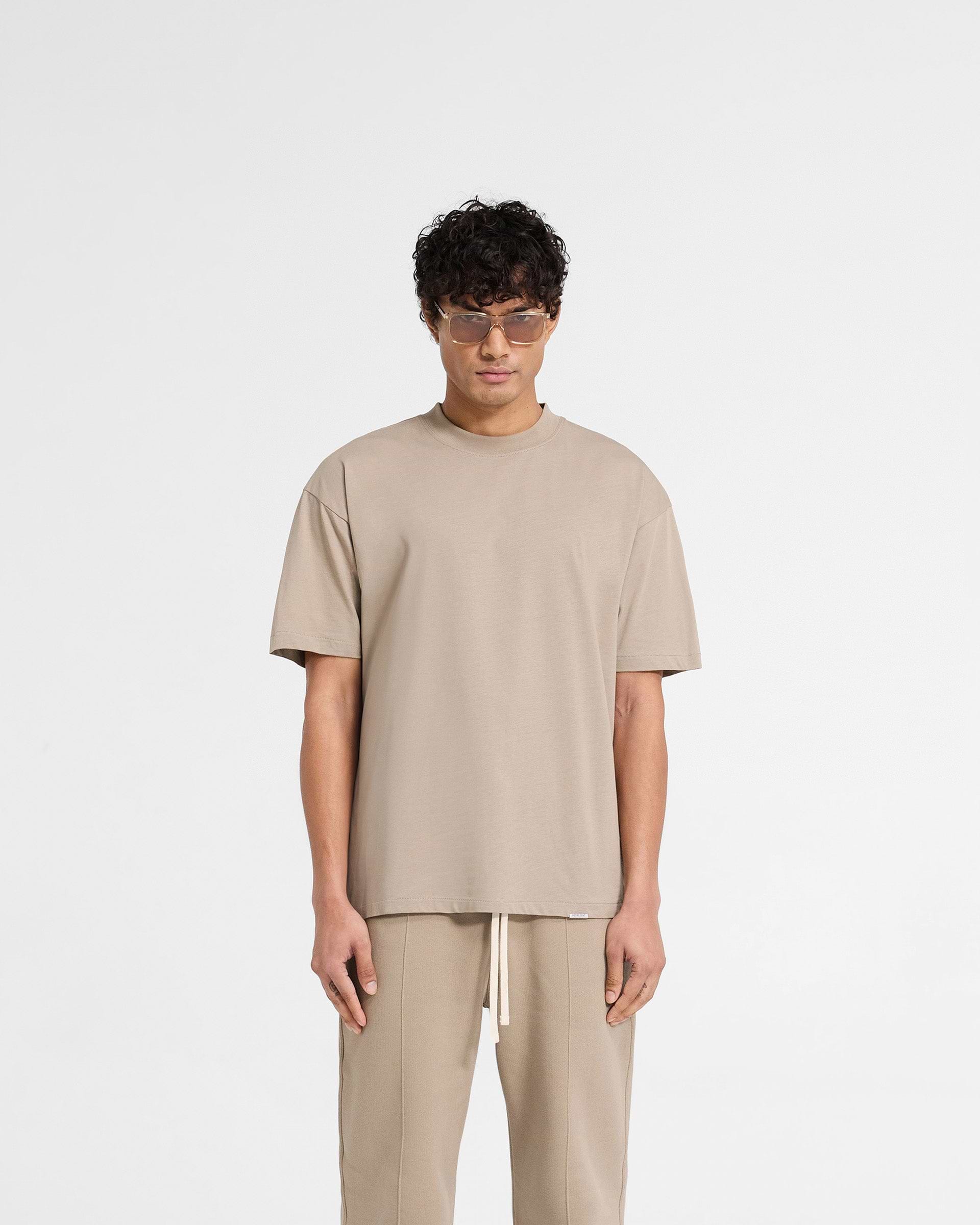 Man wearing an oversized beige t-shirt on a plain white background | Initial Oversized T-Shirt in Dune | Oversized t-shirt with embroidered details | Initial T-Shirts | Represent Clo