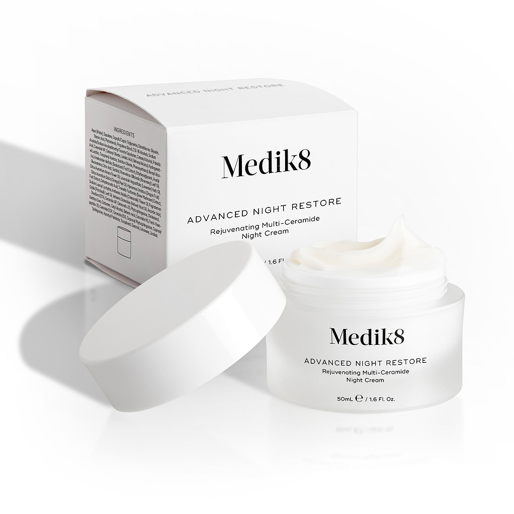 Medik8 Advanced Night Restore Cremetiegel steht offen mit dem weißen Deckel angelehnt und der Produktverpackung dahinter. Das Bild zeigt die reichhaltige Textur der Creme und das saubere Verpackungsdesign.