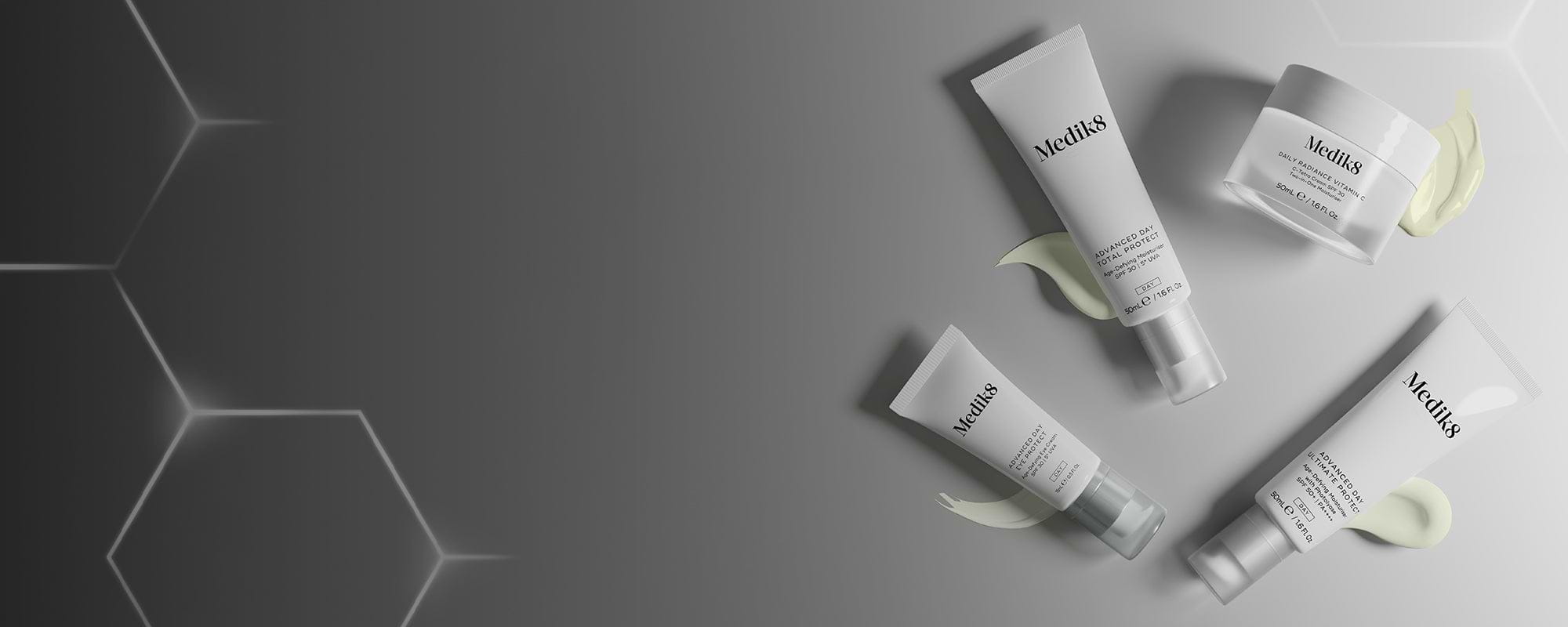 Ein Banner mit einer Anordnung weißer Medik8-Tagespflegeprodukte auf der rechten Seite. Die Produktgruppe umfasst drei Tuben – Advanced Day Total Protect, Advanced Day Eye Protect und Advanced Day Ultimate Protect – sowie einen Tiegel mit Daily Radiance Vitamin C. Jedes Produkt ist mit einem glatten, hell cremefarbenen Produktswatch darunter dargestellt. Der Hintergrund verläuft von einem dunklen Grau auf der linken Seite mit leuchtenden geometrischen Hexagonen zu einem helleren, einfarbigen Grau auf der re