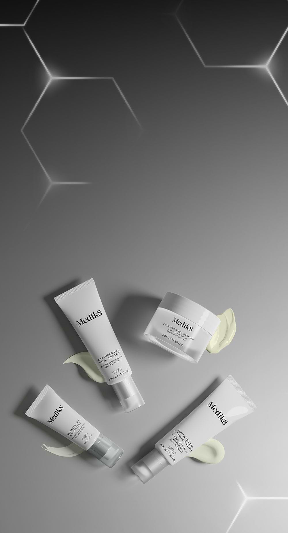 Ein Banner mit einer Anordnung weißer Medik8-Tagespflegeprodukte auf der rechten Seite. Die Produktgruppe umfasst drei Tuben – Advanced Day Total Protect, Advanced Day Eye Protect und Advanced Day Ultimate Protect – sowie einen Tiegel mit Daily Radiance Vitamin C. Jedes Produkt ist mit einem glatten, hell cremefarbenen Produktswatch darunter dargestellt. Der Hintergrund verläuft von einem dunklen Grau auf der linken Seite mit leuchtenden geometrischen Hexagonen zu einem helleren, einfarbigen Grau auf der re