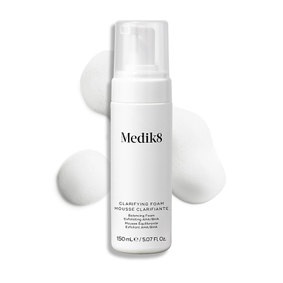 Botella con dispensador de Medik8 Clarifying Foam exhibida sobre un fondo blanco limpio junto a tres porciones de espuma blanca y ligera. El texto identifica el producto como un limpiador refrescante para imperfecciones con AHA/BHA de 150 mL.