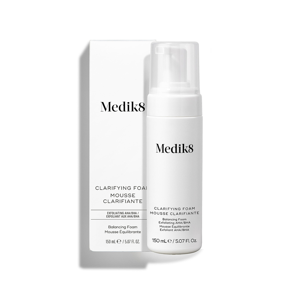 La botella con dispensador de Medik8 Clarifying Foam se encuentra junto a su caja exterior blanca a juego sobre un fondo blanco limpio. El texto en ambos identifica el producto como un limpiador refrescante para imperfecciones con AHA/BHA de 150 mL.