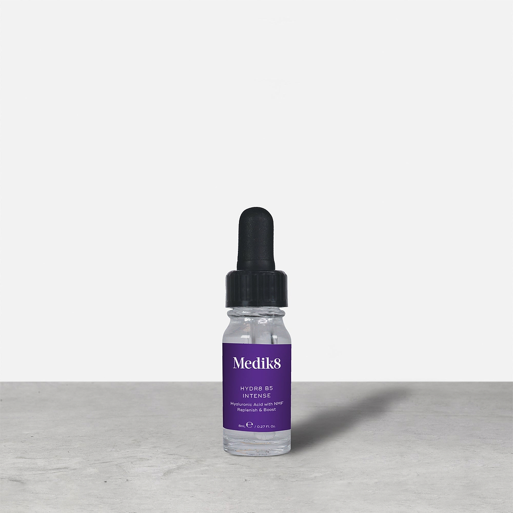 Botella de vidrio con gotero de 8 ml de Medik8 HYDR8 B5 INTENSE en color púrpura colocada junto a su caja de embalaje blanca sobre una superficie gris con fondo blanco. El texto en ambos indica: «Medik8, HYDR8 B5 INTENSE, Ácido hialurónico con NMF Replenish & Boost, 8 ml / 0,27 fl. oz.».
