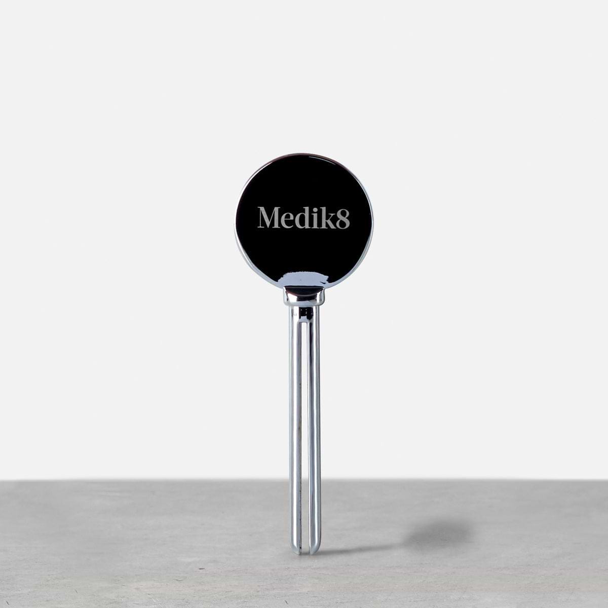 Un exprimidor metálico plateado de tubos de Medik8 en posición vertical sobre una superficie gris. El cabezal circular presenta el logotipo de Medik8, diseñado para ayudar a dispensar el producto de forma eficiente.