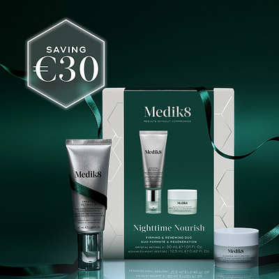 A Medik8 "Nighttime Nourish" gift box wrapped with a dark green ribbon on a reflective surface against a dark green background. The white box features a dark green center panel displaying the printed products inside: a Crystal Retinal 3 tube and an Advanced Night Restore travel size jar. Text highlights "Firming & Renewing Duo."