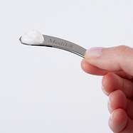 A hand holds a small, curved silver metal spatula with the "Medik8" logo engraved. A small dollop of white cream is visible on the tip of the spatula