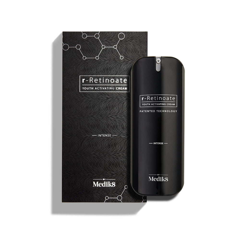 Le flacon de Medik8 r-Retinoate Intense Youth Activating Cream est placé à côté de son emballage extérieur noir, avec des graphiques hexagonaux chimiques blancs et un motif ondulé texturé 