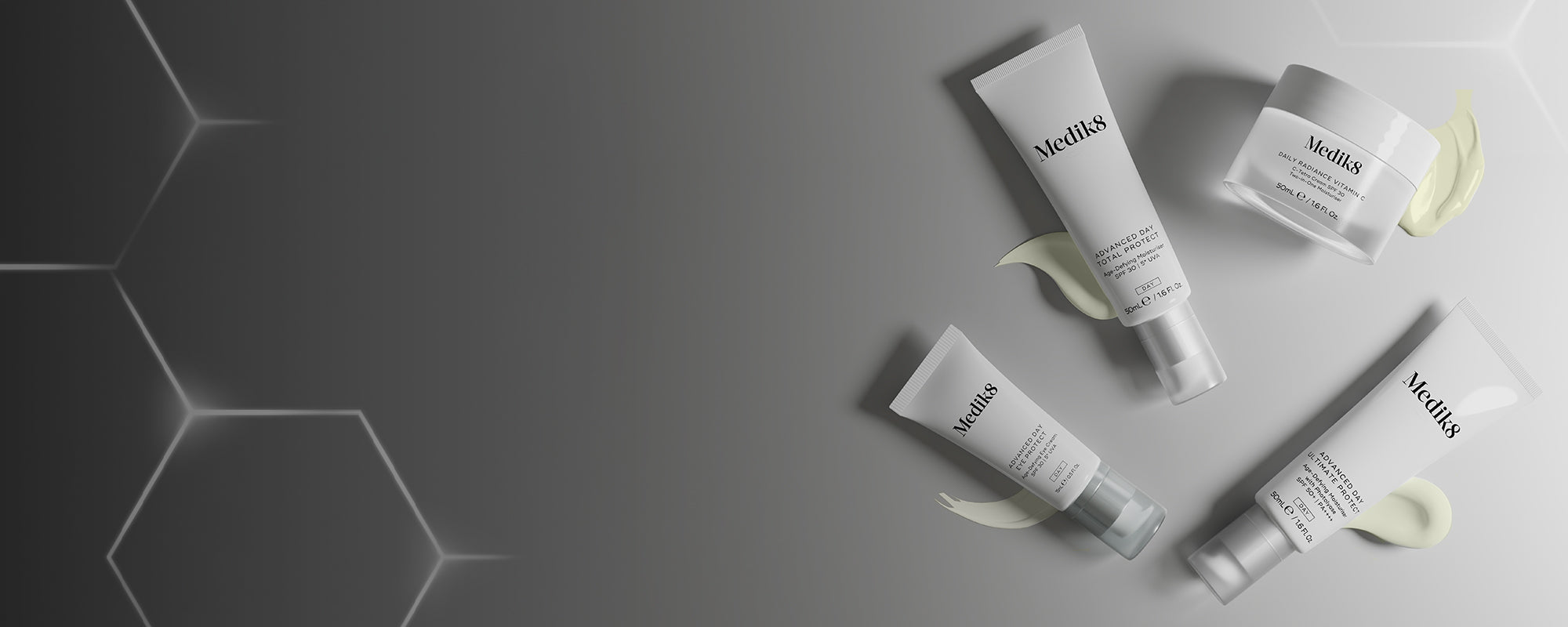 Une bannière présentant une sélection de soins de jour Medik8 blancs disposés sur le côté droit. L’ensemble comprend trois tubes — Advanced Day Total Protect, Advanced Day Eye Protect et Advanced Day Ultimate Protect — ainsi qu’un pot de Daily Radiance Vitamin C. Chaque produit est associé à une texture de produit lisse dans une teinte crème claire située en dessous. L’arrière-plan évolue d’un gris foncé à gauche, avec des hexagones géométriques lumineux, vers un gris uni plus clair à droite.