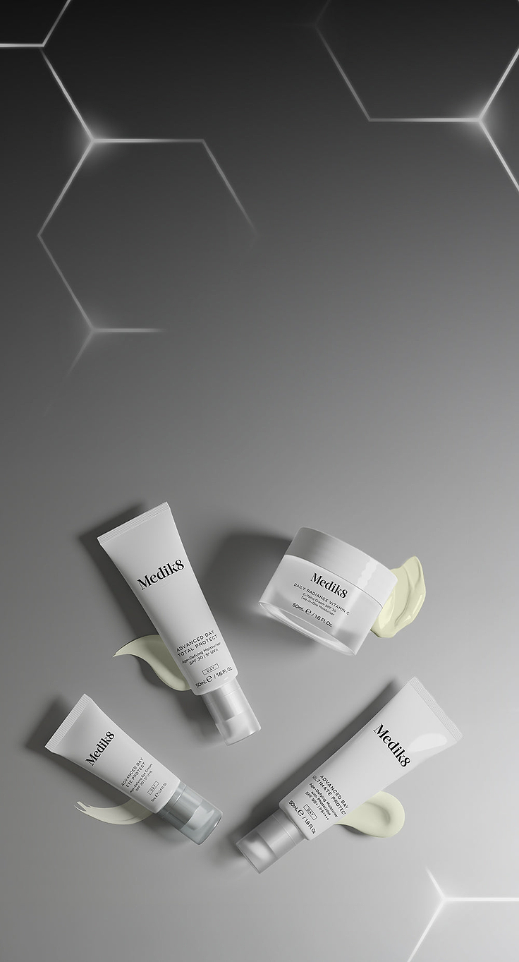 Une bannière présentant une sélection de soins de jour Medik8 blancs disposés sur le côté droit. L’ensemble comprend trois tubes — Advanced Day Total Protect, Advanced Day Eye Protect et Advanced Day Ultimate Protect — ainsi qu’un pot de Daily Radiance Vitamin C. Chaque produit est associé à une texture de produit lisse dans une teinte crème claire située en dessous. L’arrière-plan évolue d’un gris foncé à gauche, avec des hexagones géométriques lumineux, vers un gris uni plus clair à droite.