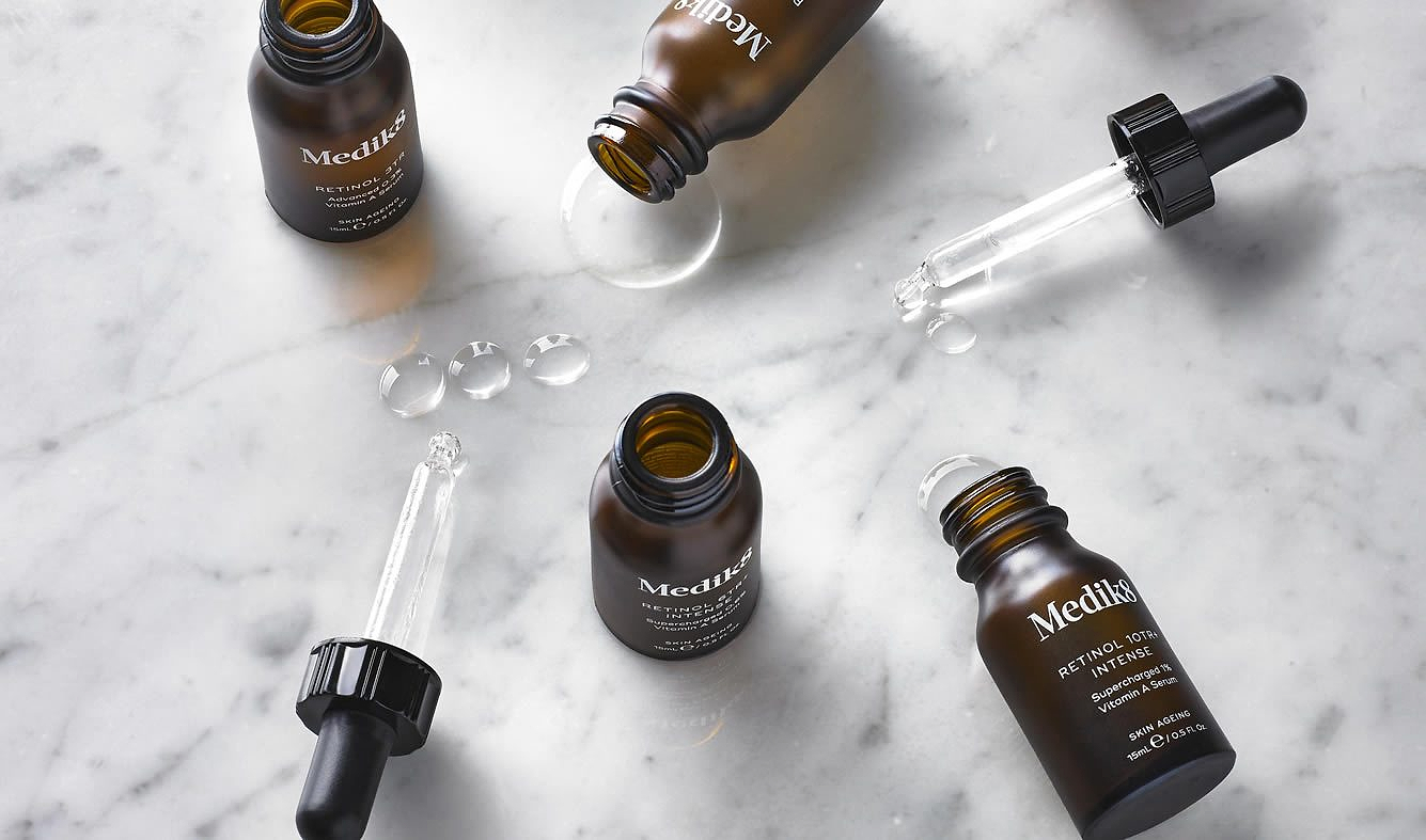A flat lay arrangement of Intelligent Retinol serums on a white marble surface. Several amber glass bottles are scattered alongside glass droppers that have dispensed clear, silky droplets of the Vitamin A serum. The composition highlights the professional, apothecary-inspired aesthetic of the 