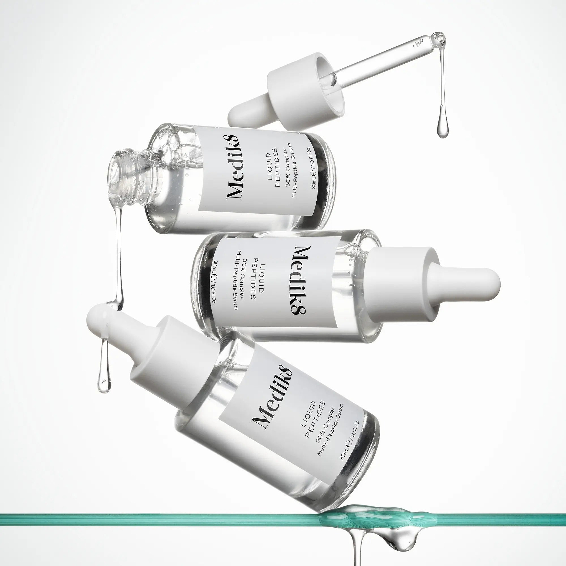 Tři lahvičky séra Medik8 Liquid Peptides 30% Complex vznášející se nad skleněným povrchem s kapkami séra.