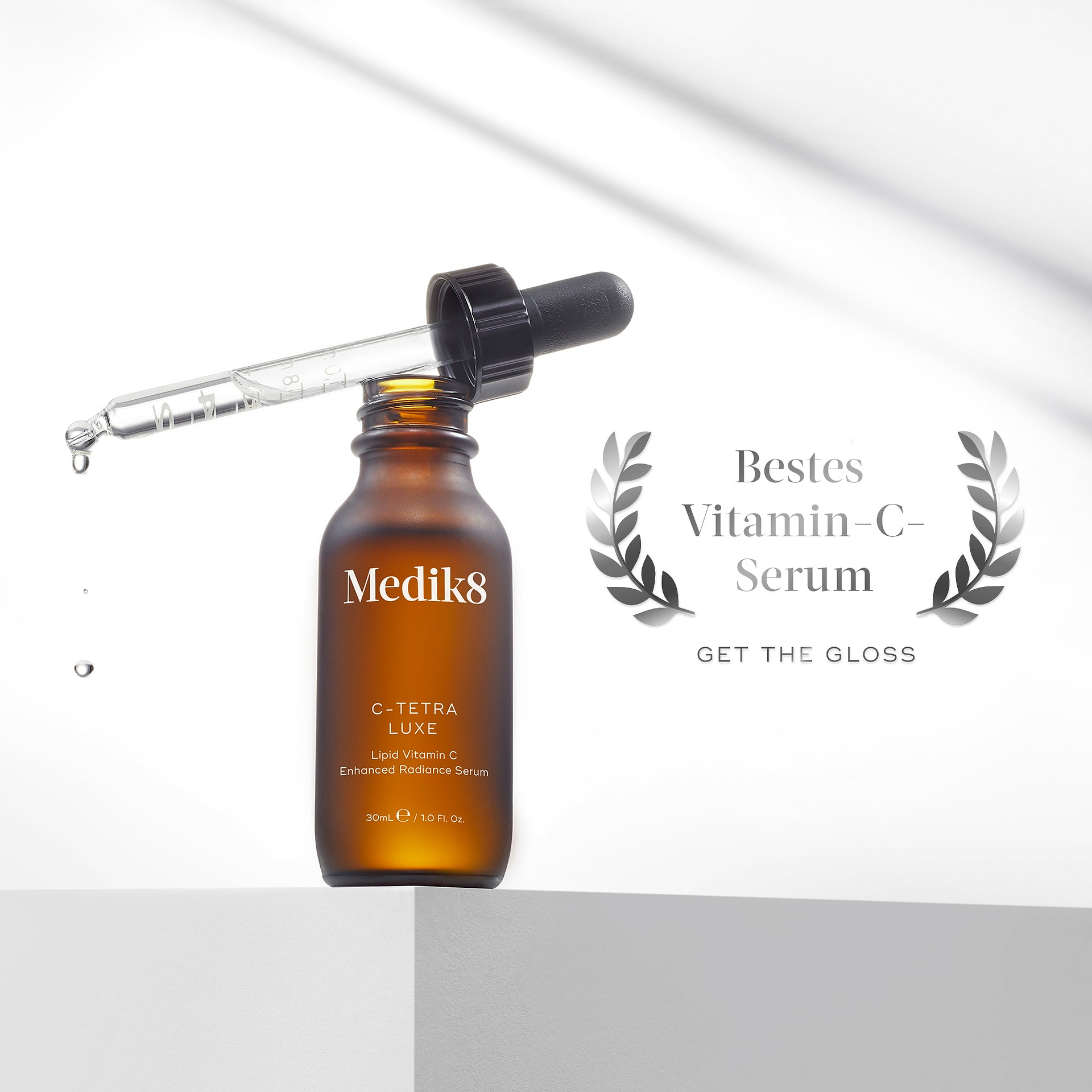 Eine Braunglasflasche des Medik8 C-Tetra Luxe Lipid Vitamin C Serums auf einem weißen Sockel. Die Pipette liegt obenauf und lässt Tropfen herabsinken. Daneben befindet sich ein silberner Lorbeerkranz mit dem Text „Bestes Vitamin-C-Serum - GET THE GLOSS“.