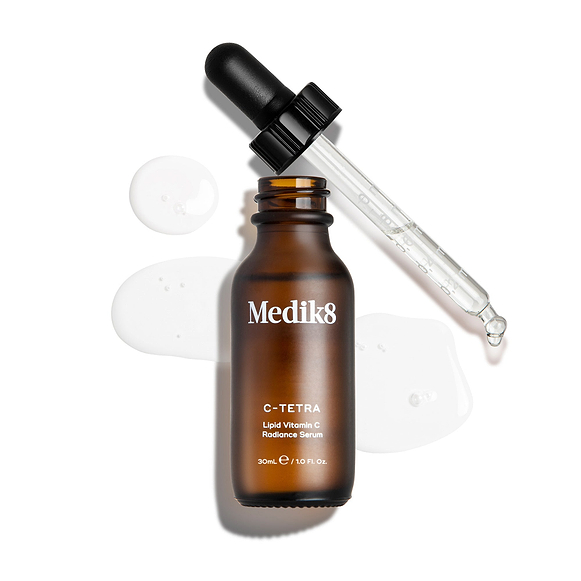 Eine Draufsicht auf eine geöffnete 30-ml-Braunglasflasche des Medik8 C-Tetra Serums. Die Glaspipette liegt daneben, umgeben von klaren, flüssigen Tropfen des Serums auf weißem Hintergrund.-6