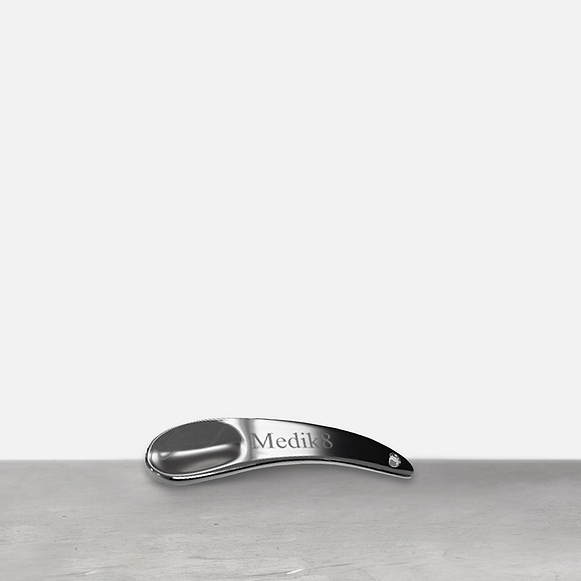Une spatule en métal argenté repose sur une surface grise. La spatule courbée porte le logo "Medik8" gravé sur son manche.-46