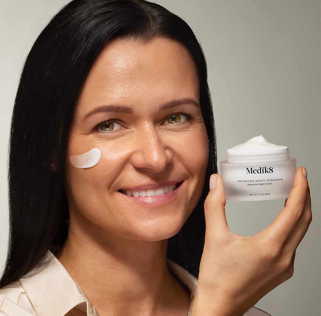 Usmievajúca sa tmavovlasá žena drží otvorenú nádobku krému Medik8 Advanced Night Ceramide Signature Night Cream.