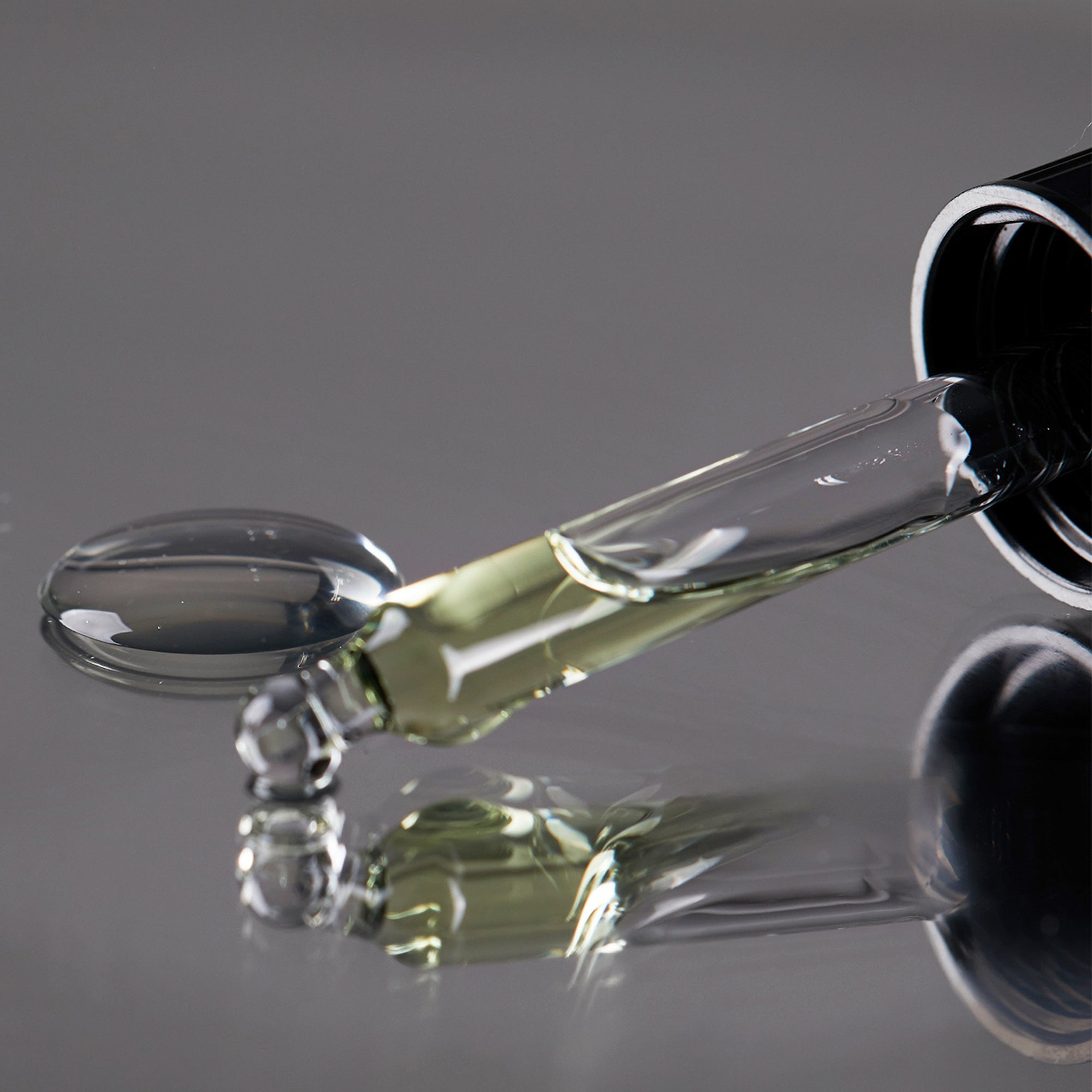 A macro close-up of a glass dropper pipette resting on a reflective silver surface. The pipette is filled with a pale yellow liquid serum, and a single, perfectly clear droplet has pooled on the surface behind it. Both the pipette and the pool of liquid cast sharp, dark reflections against the metallic background, emphasizing a clean and clinical aesthetic.