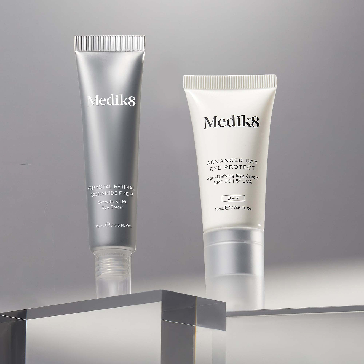 Two Medik8 eye care tubes stand on clear acrylic blocks against a soft grey background. On the left is the silver Crystal Retinal Ceramide Eye 6 (for night use) and on the right is the white Advanced Day Eye Protect SPF 30 (for day use), showcasing a complete 24-hour eye care routine.