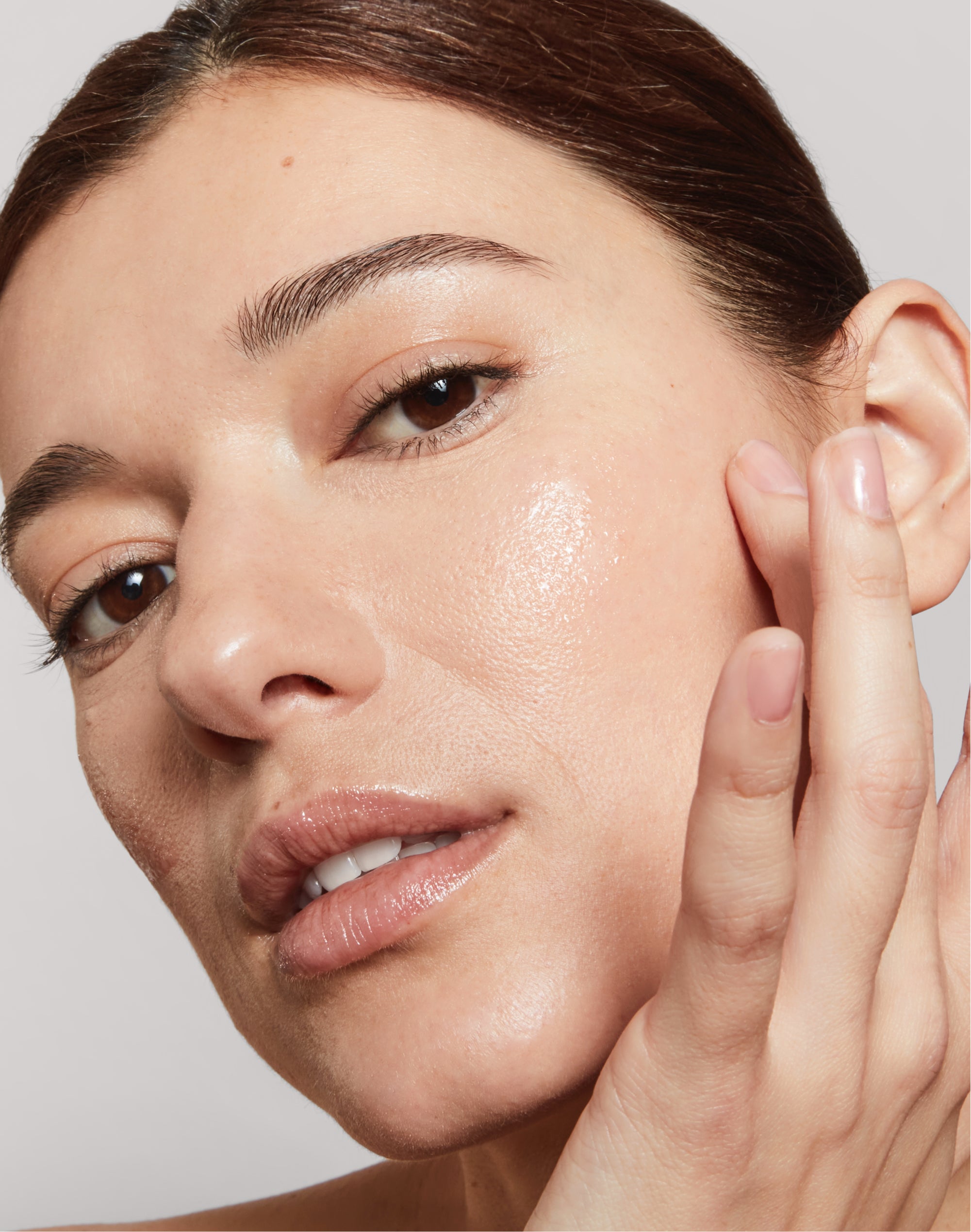 An extreme close-up of a woman with her hair pulled back, showcasing a radiant and dewy skin texture. She has her hand raised to her cheek, gently touching her face with her fingertips. The lighting highlights the natural glow and smooth finish of her complexion, emphasizing a healthy and hydrated appearance.