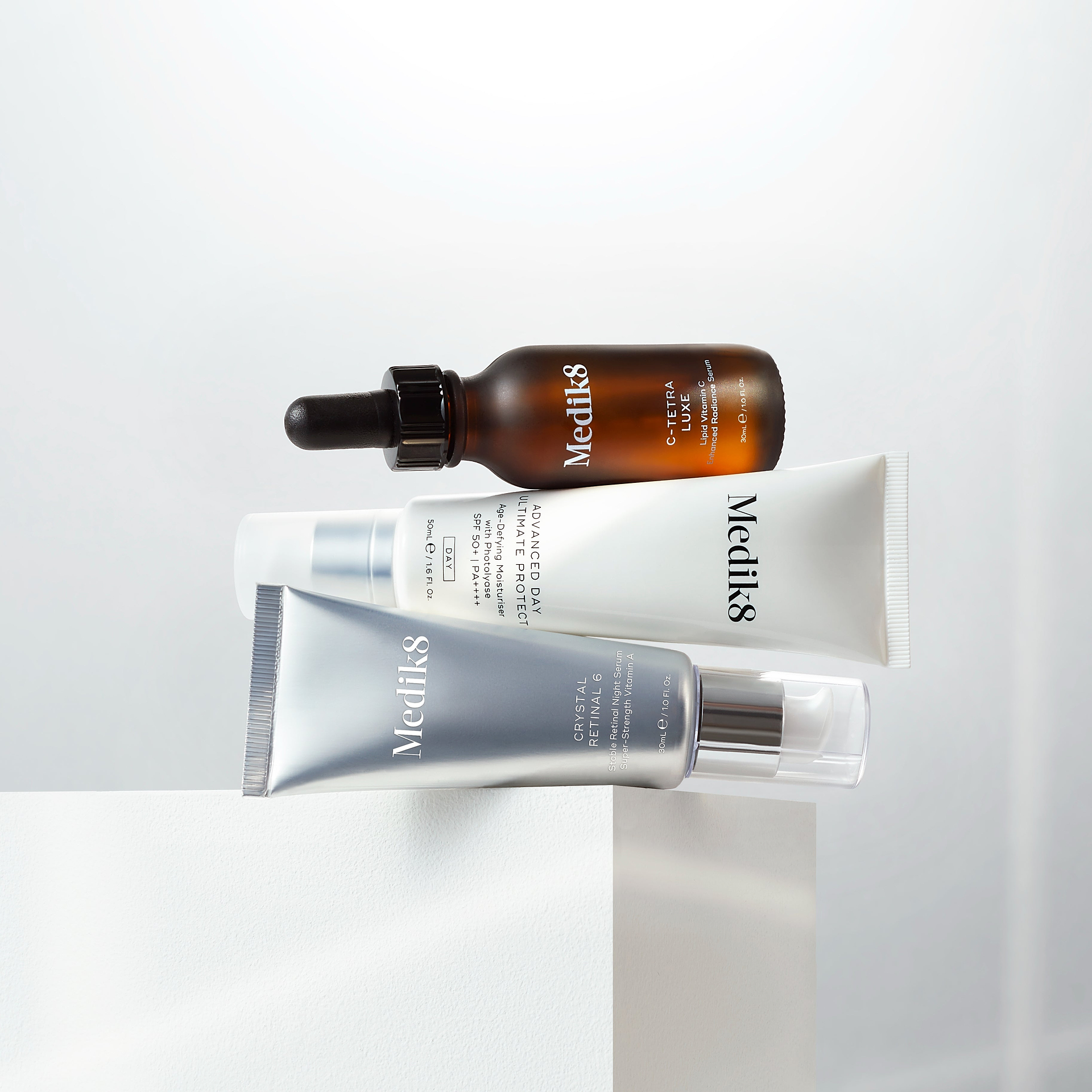 A stack of Medik8 skincare products balanced on a white block against a white background. From top to bottom: C-Tetra Luxe serum bottle (Liquid Vitamin C), Advanced Day Ultimate Protect tube (Age-Defying Moisturiser with SPF 50+), and Crystal Retinal 6 tube (Stable Retinal Night Serum).