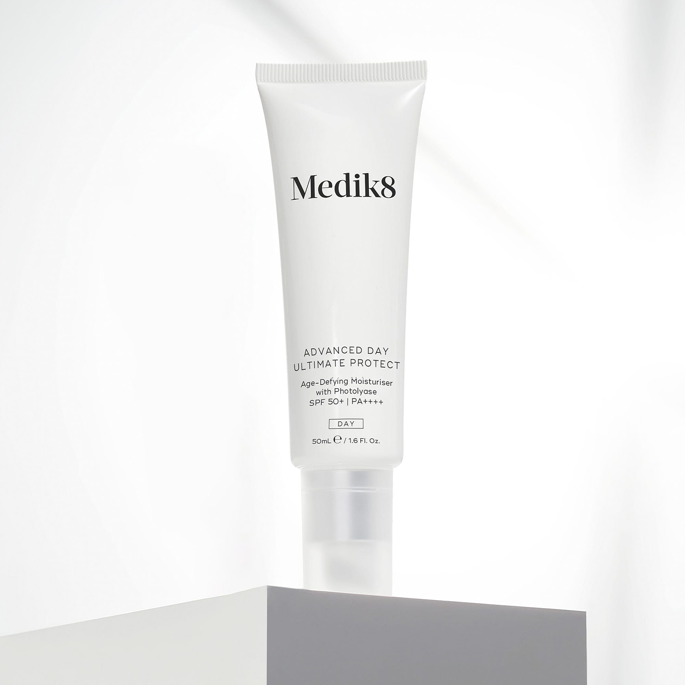 A white 50mL tube of Medik8 Advanced Day Ultimate Protect cream standing upright on a light grey geometric block. Set against a soft, draped white fabric background, the text on the tube highlights it as an 