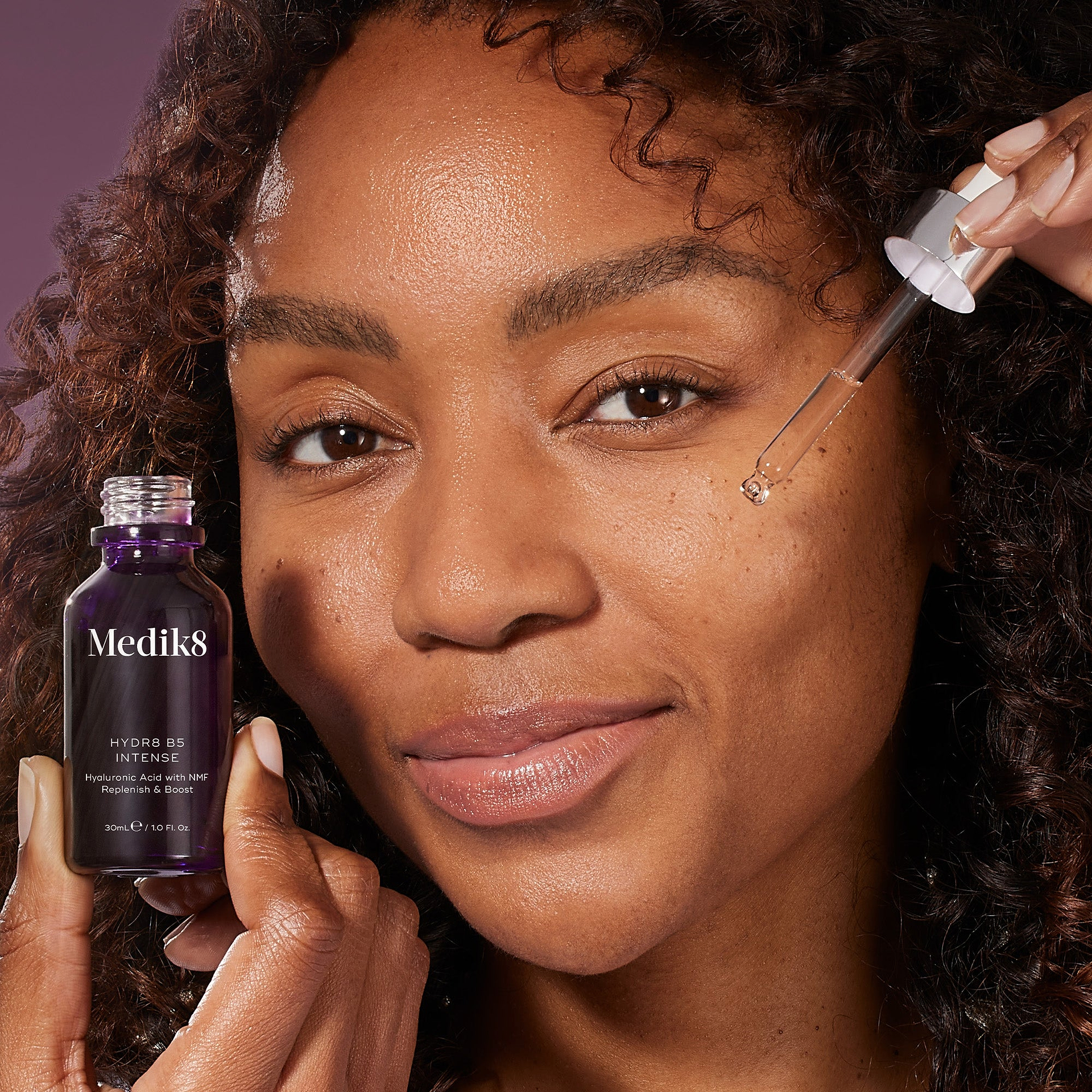 A woman with curly hair smiles while holding a Medik8 Hydr8 B5 Intense serum bottle and a glass dropper near her face. The purple bottle features text for Hyaluronic Acid with NMF, Replenish & Boost, set against a deep purple background.