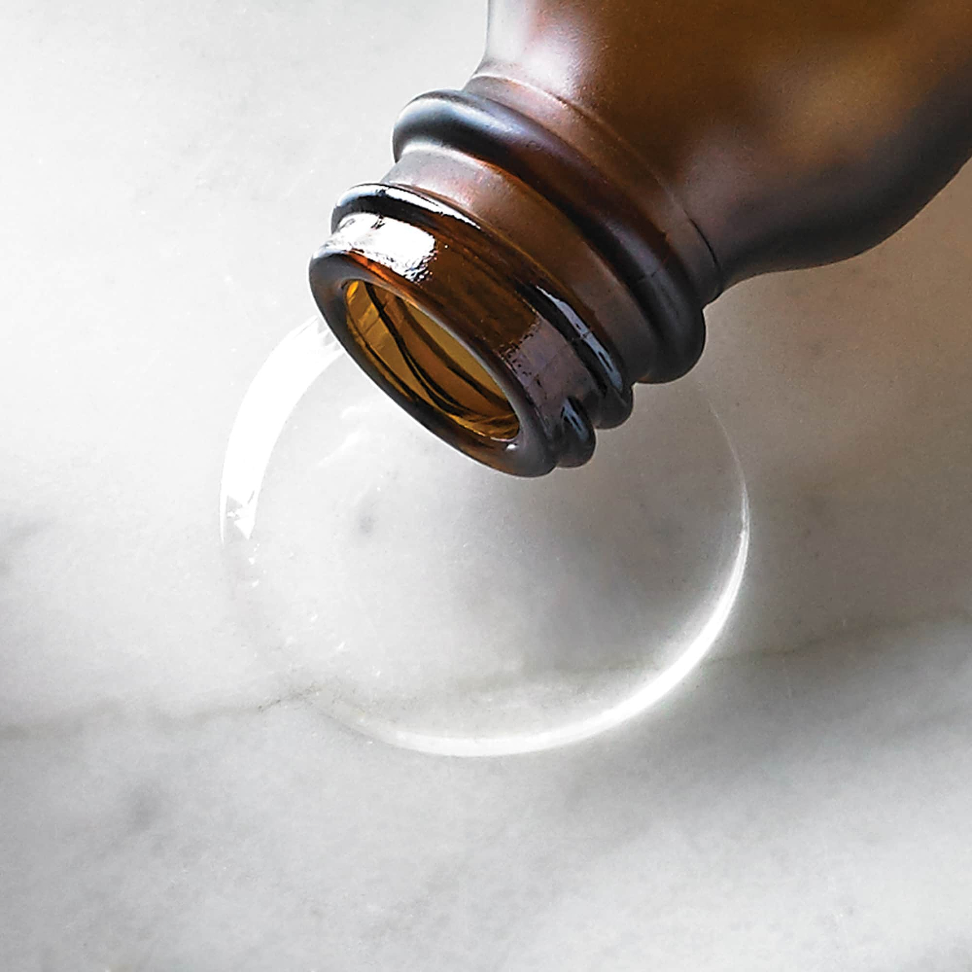 Close-up of an amber Intelligent Retinol Eye TR serum bottle neck as it dispenses a clear, liquid droplet onto a white marble surface.