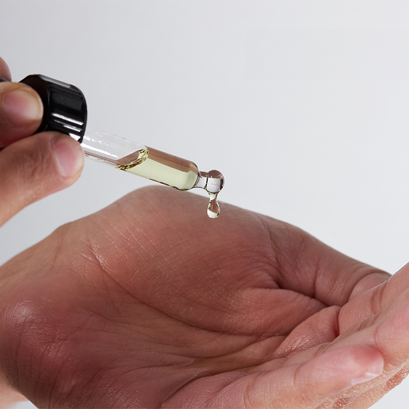 Close-up of a person’s palm as a glass dropper dispenses a single drop of pale yellow oil-based serum, highlighting the liquid texture against a clean white background.