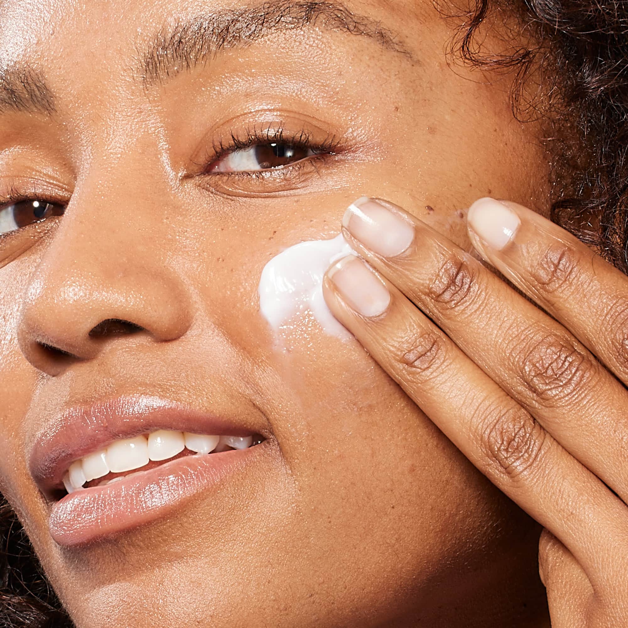 Close-up of a smiling woman with a radiant complexion applying a swipe of white Medik8 Total Moisture Daily Facial Cream to her cheek with her fingers, demonstrating how to use the product.