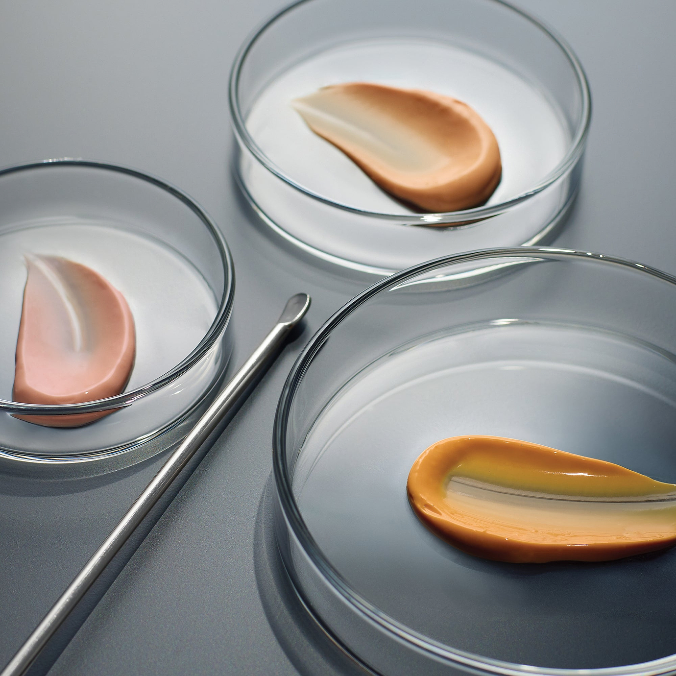 A clinical, high-angle shot of three clear glass petri dishes placed on a slate-grey surface. Each dish contains a smooth, swept swatch of Crystal Retinal serum in varying shades of peach and orange, demonstrating the color change associated with increasing concentrations of retinaldehyde. A stainless steel lab spatula lies beside the dishes, emphasizing the brand's professional and scientific heritage.
