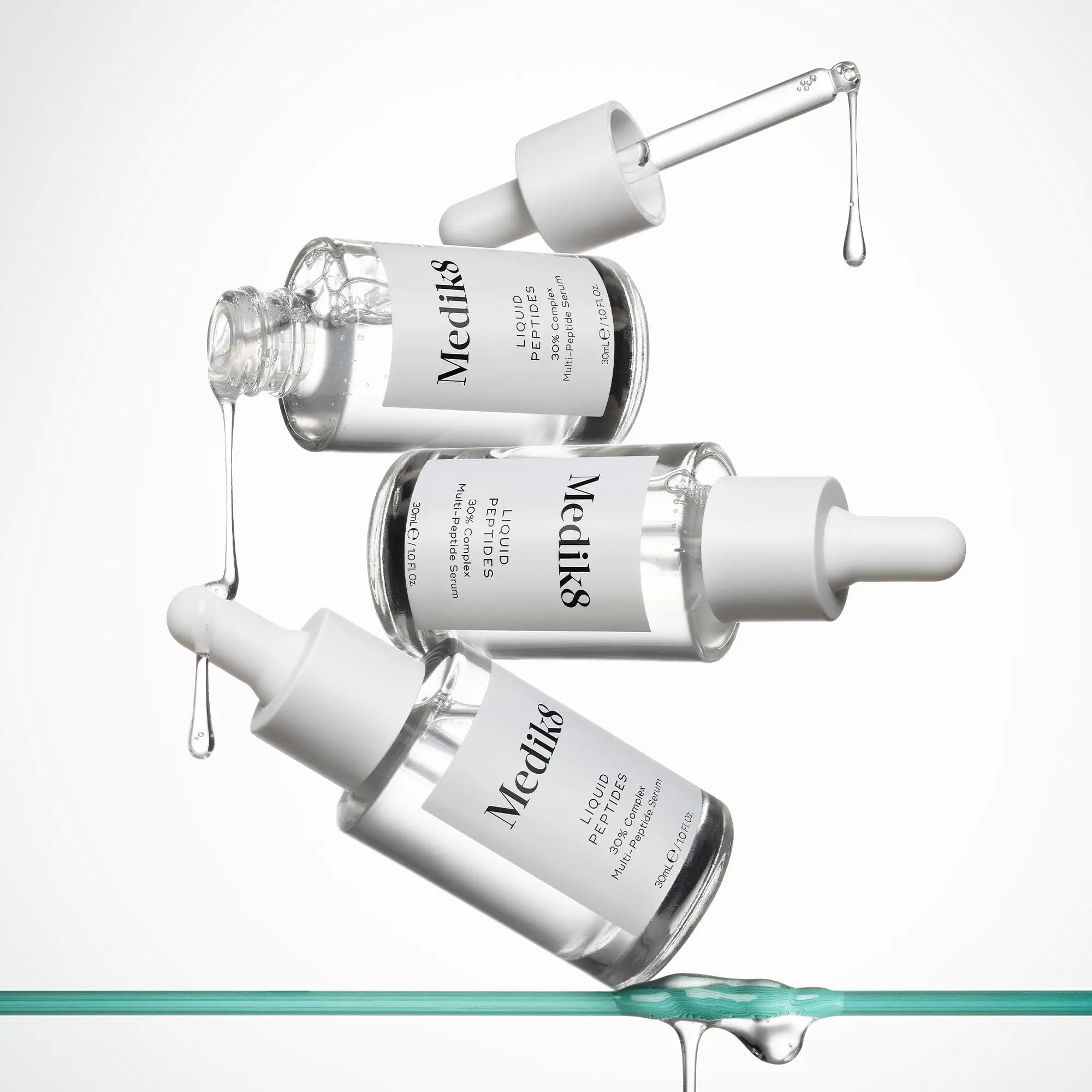 An artistic, high-key composition featuring three clear glass bottles of Liquid Peptides 30% Complex Multi-Peptide Serum. The bottles appear to be floating or falling against a pure white background, with clear serum droplets suspended in mid-air and dripping from the pipettes, conveying a sense of hydration and fluidity.