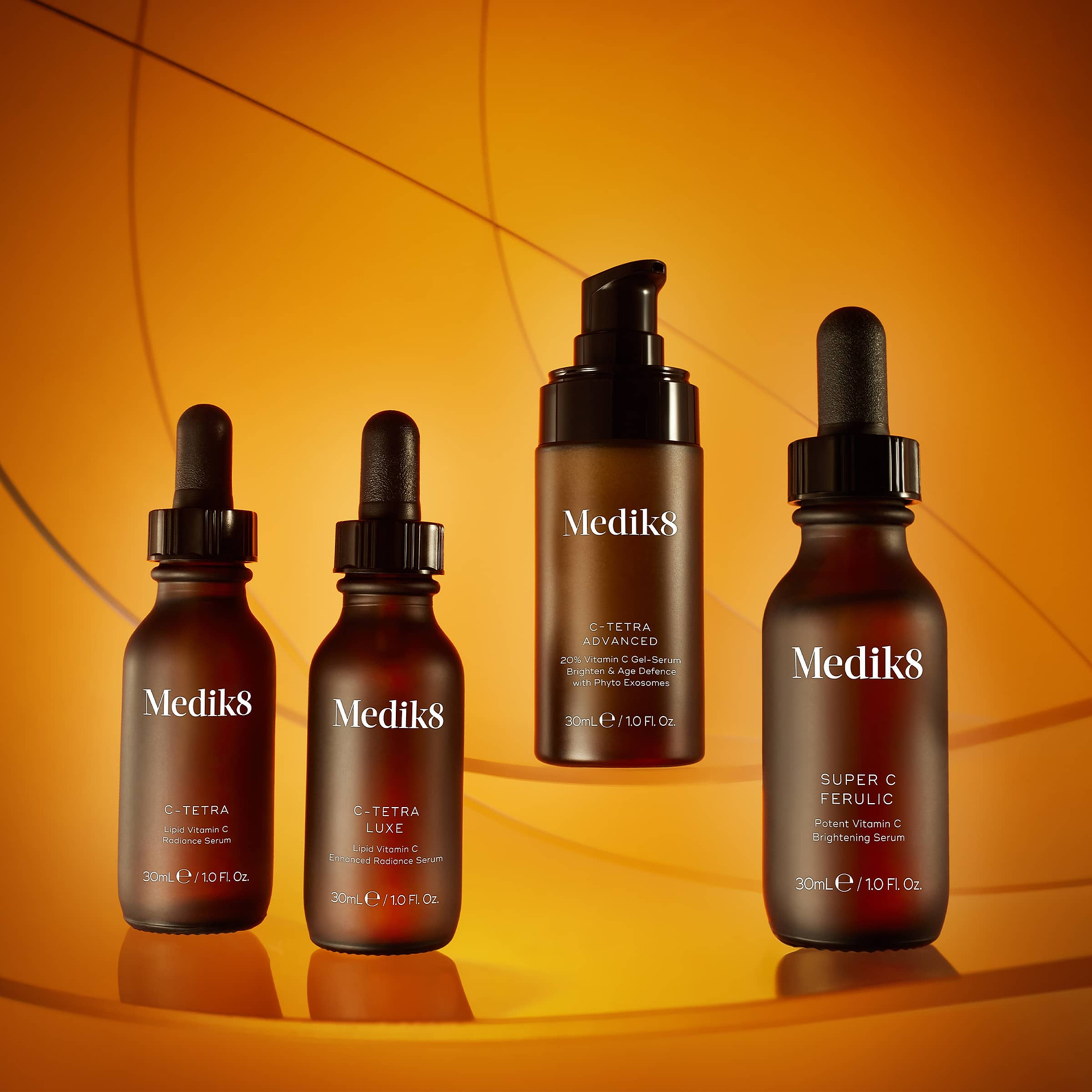 Four Medik8 Vitamin C serums displayed against a warm amber background with geometric lines. From left to right: C-Tetra (Liquid Vitamin C Radiance Serum), C-Tetra Luxe (Enhanced Radiance Serum), C-Tetra Advanced (20% Vitamin C Gel-Serum), and Super C Ferulic (Potent Vitamin C Brightening Serum).