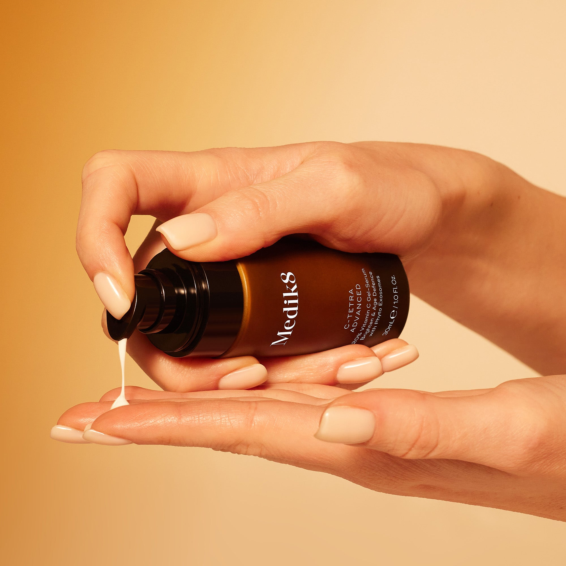 Close-up of a person's hands dispensing Medik8 C-Tetra Advanced gel-serum from an amber pump bottle onto their fingers. Set against a warm orange background, the image highlights the product's white, creamy texture as it is being pumped out.