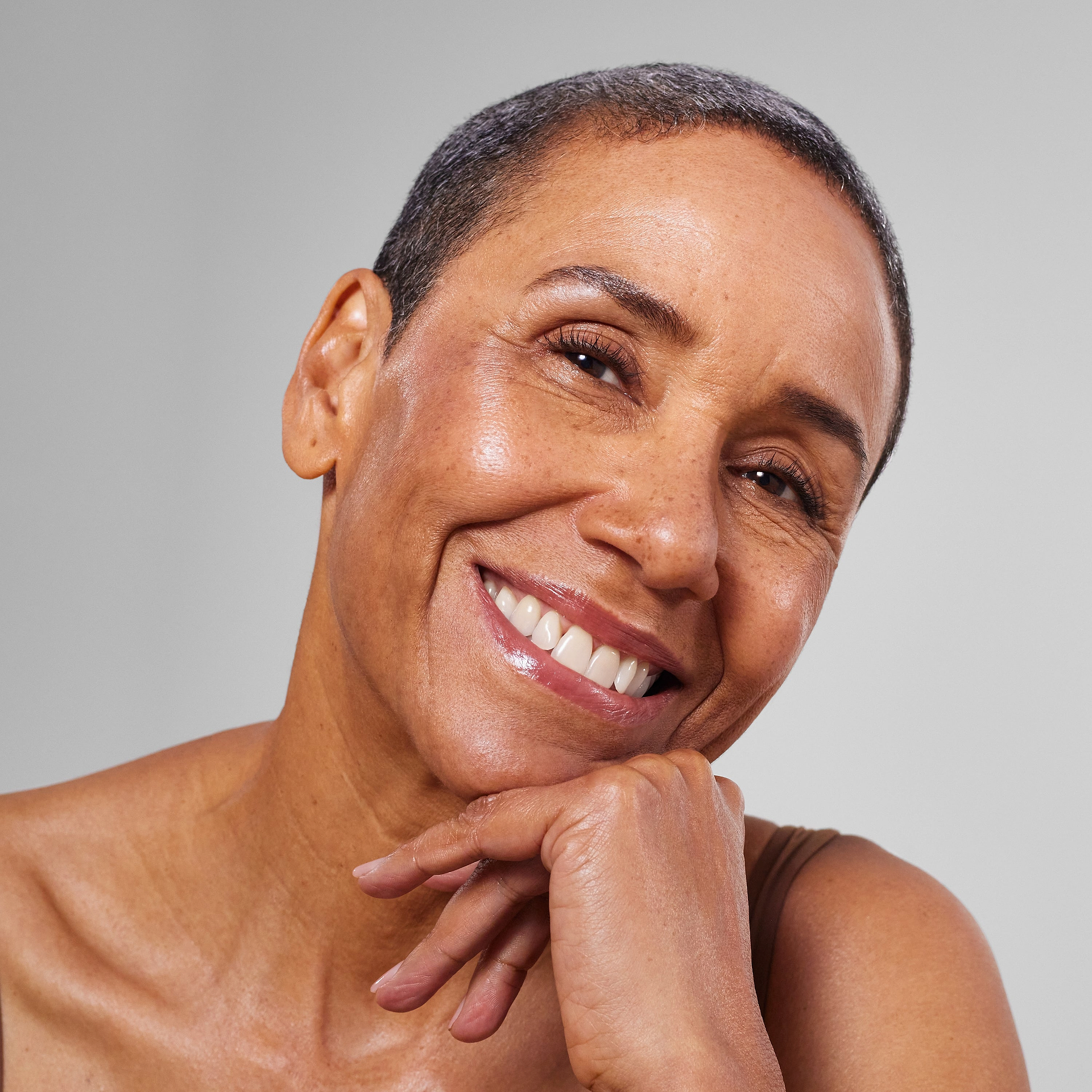 Close-up portrait of a smiling woman with short hair and a glowing, radiant complexion, resting her chin on her hand against a neutral light grey background.