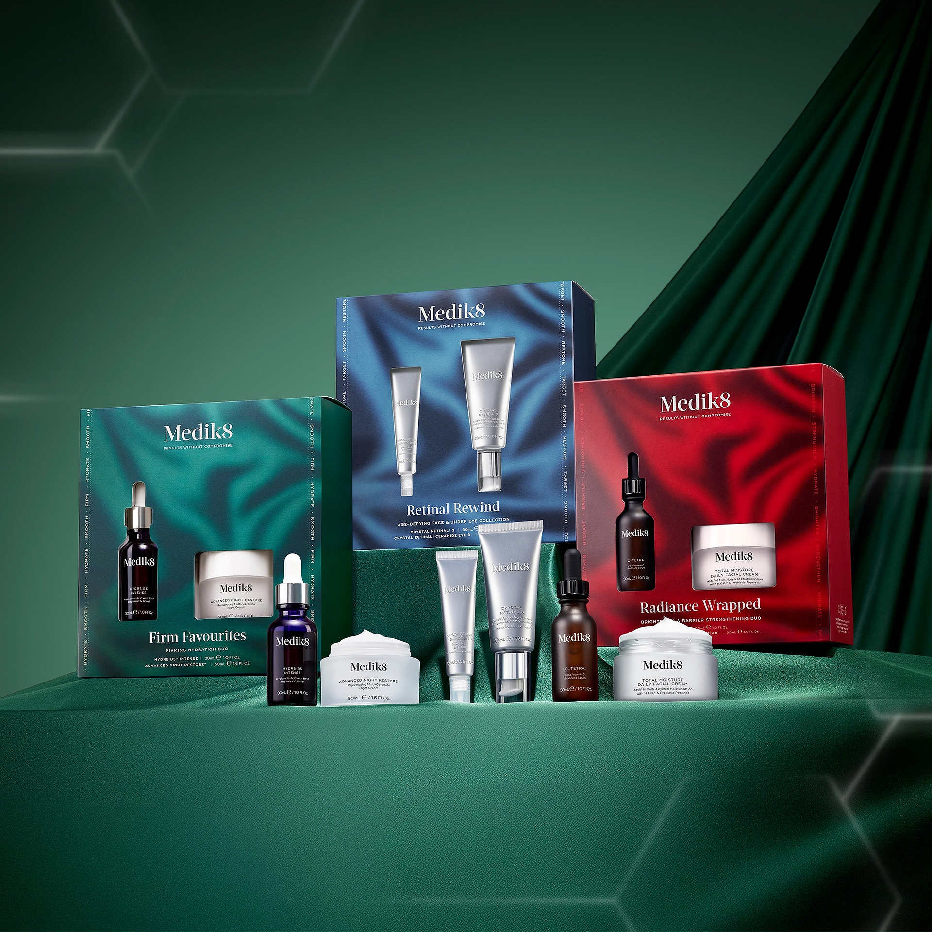 A collection of three festive Medik8 gift sets: Firm Favourites (teal), Retinal Rewind (blue), and Radiance Wrapped (red). The individual products from each set, including various serums and moisturizers, are lined up in front of their respective colorful boxes. The scene is set on a forest green draped surface with faint white geometric patterns in the background, creating a professional holiday display.